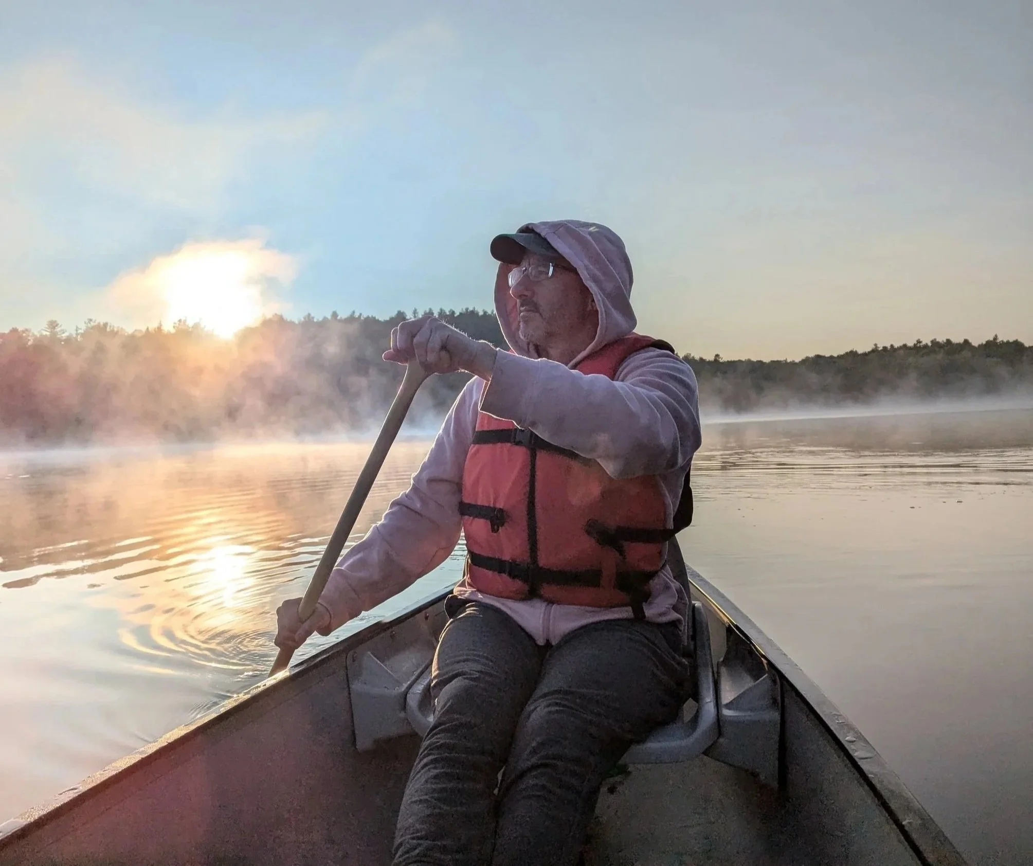 Man in a boat on a river at sunset, wearing a hoodie and life jacket, paddling with a paddle, with mist on the water and trees in the background.
