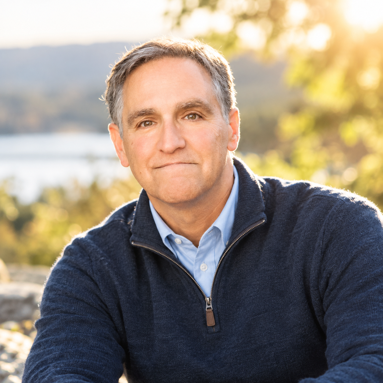 A middle-aged man with gray hair sitting outdoors in a natural setting, wearing a navy blue zip-up sweater over a light blue collared shirt, during golden hour sunlight.
