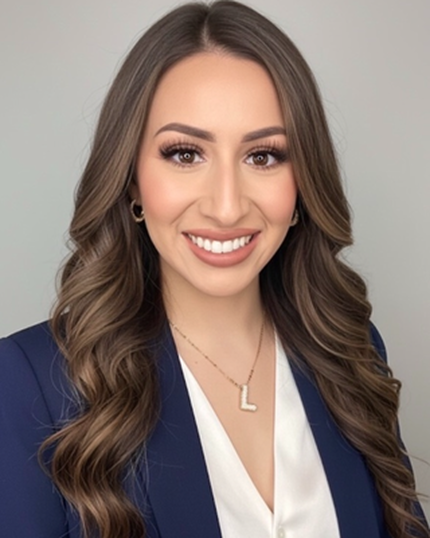 A young woman with long, wavy brown hair, smiling, wearing a navy blazer, a white blouse, gold earrings, and a necklace with a letter 'L', posed against a plain gray background.