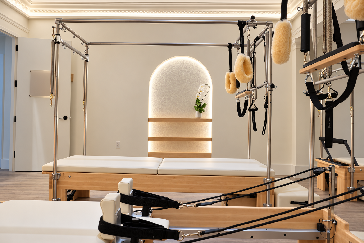 A modern physical therapy or rehabilitation room with specialized equipment, including a wooden therapy bed, suspension and pulley systems, and padded supports, with a decorative orchid on a backlit wall and neutral-colored walls.