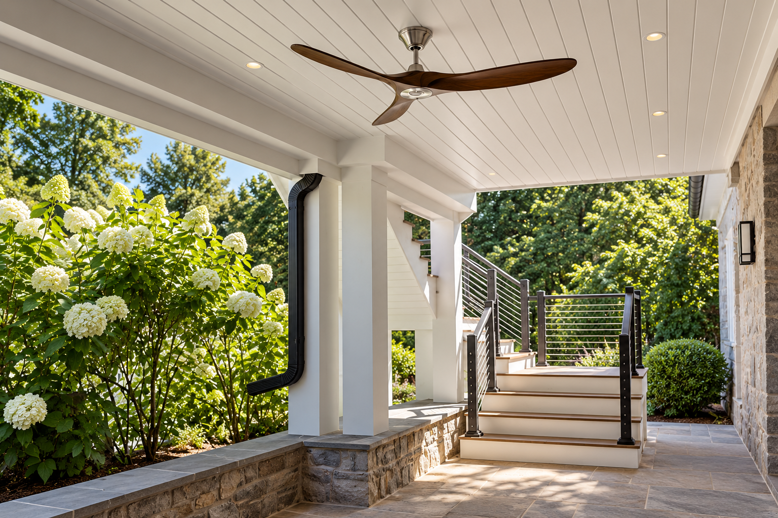 Clean and modern outdoor porch area with a ceiling fan, stairs, and a black railing, surrounded by green foliage and white flowers.