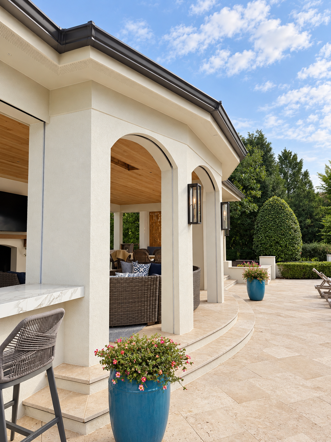 A spacious outdoor patio with arched openings, wicker chairs with blue and white cushions, potted flowering plants in blue pots, and a view of a well-manicured garden with tall trees under a partly cloudy sky.