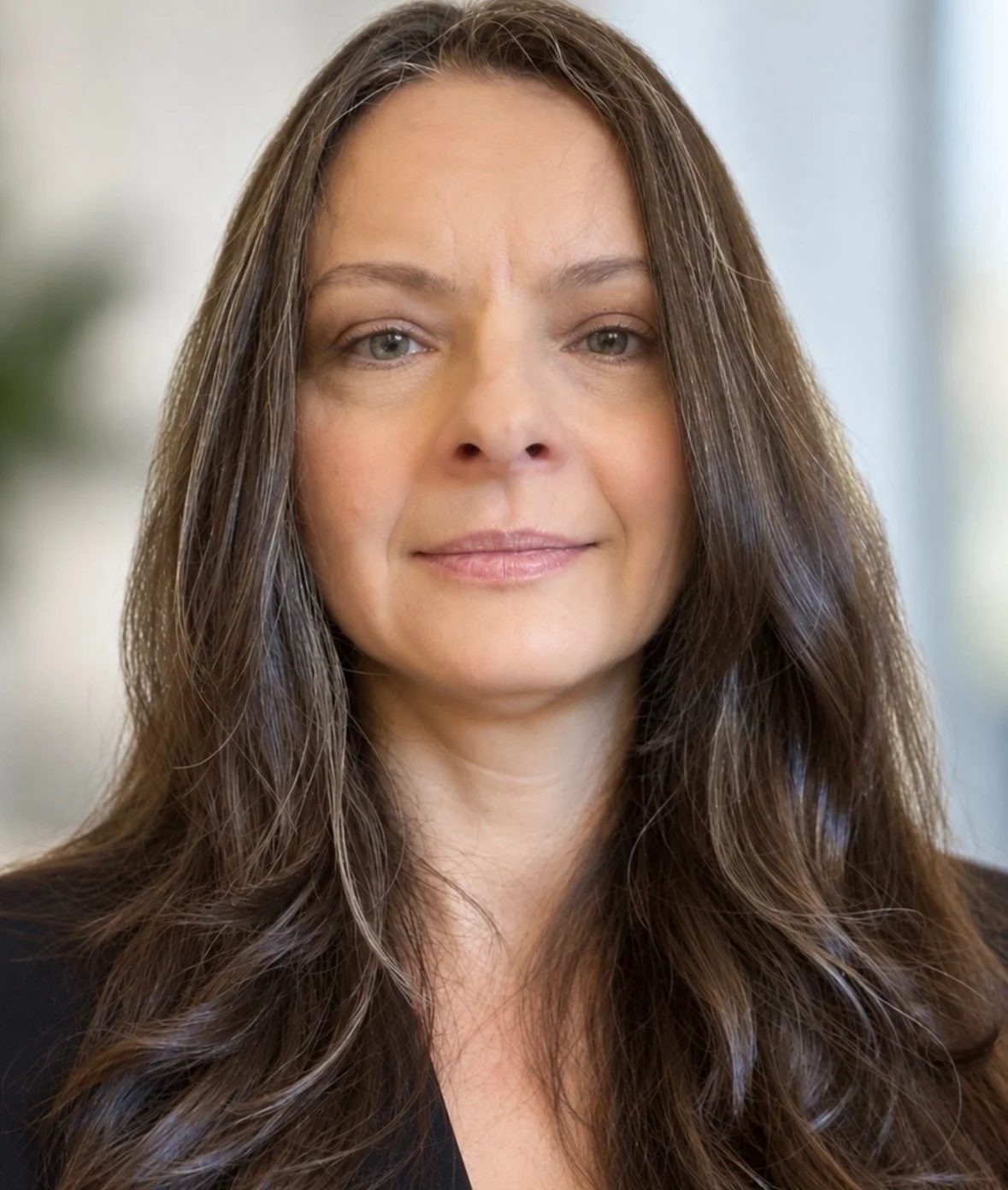 Close-up portrait of a woman with long, wavy brown hair, fair skin, and light-colored eyes, wearing a dark top with a blurred indoor background.