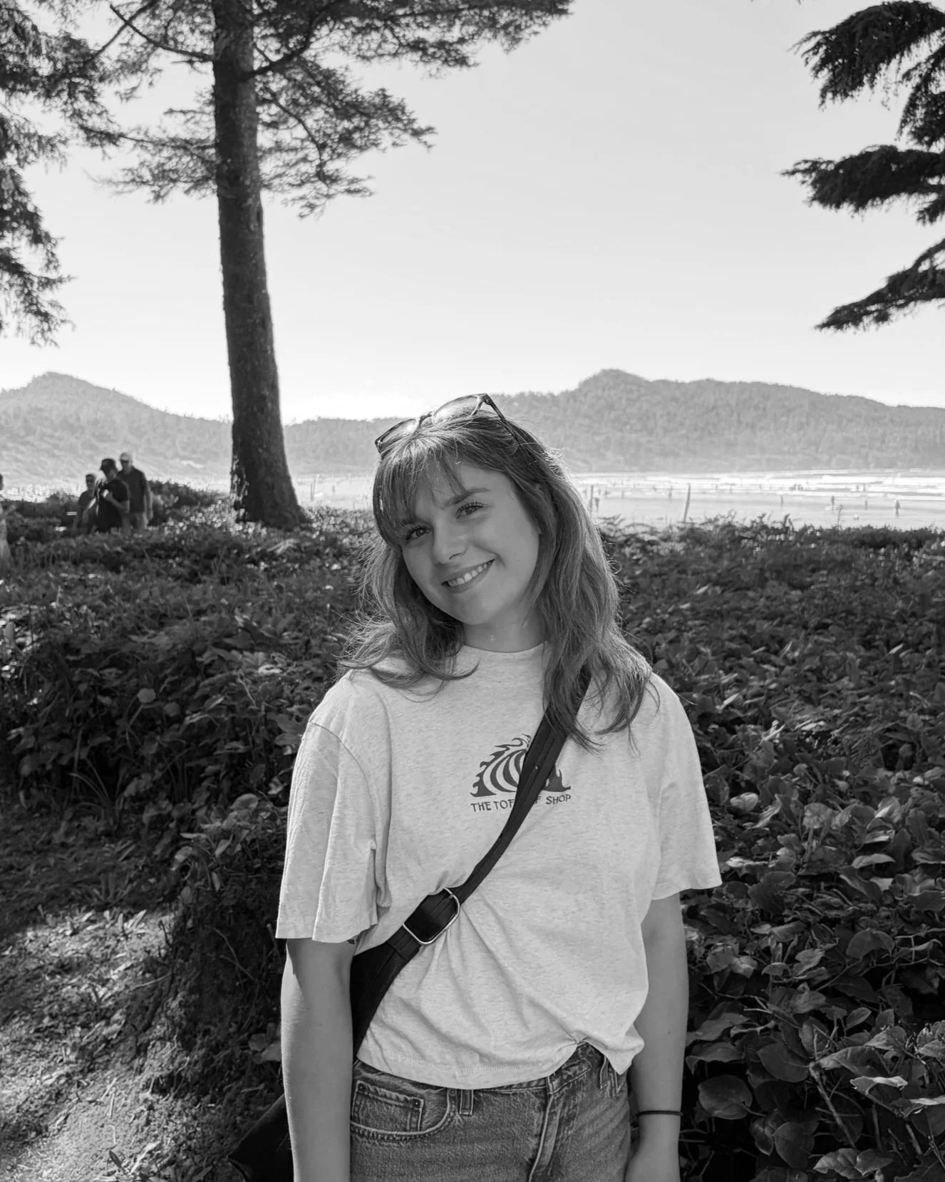 A smiling young woman with shoulder-length hair, wearing sunglasses on her head, a T-shirt, and jeans, standing outdoors among trees and plants, with mountains and a body of water in the background.