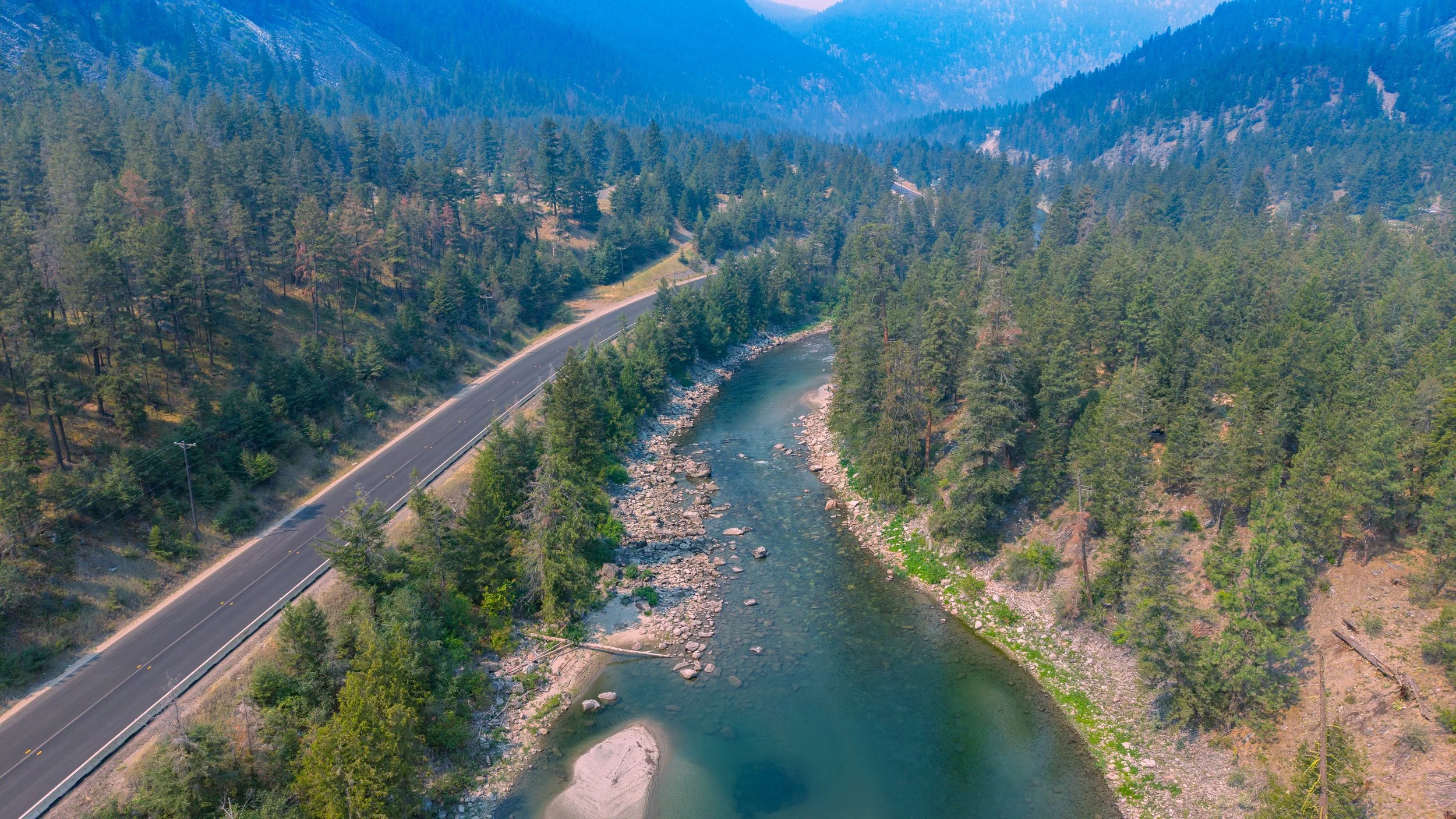 Aerial view of a winding mountain road running alongside a river through a dense forest of pine trees.