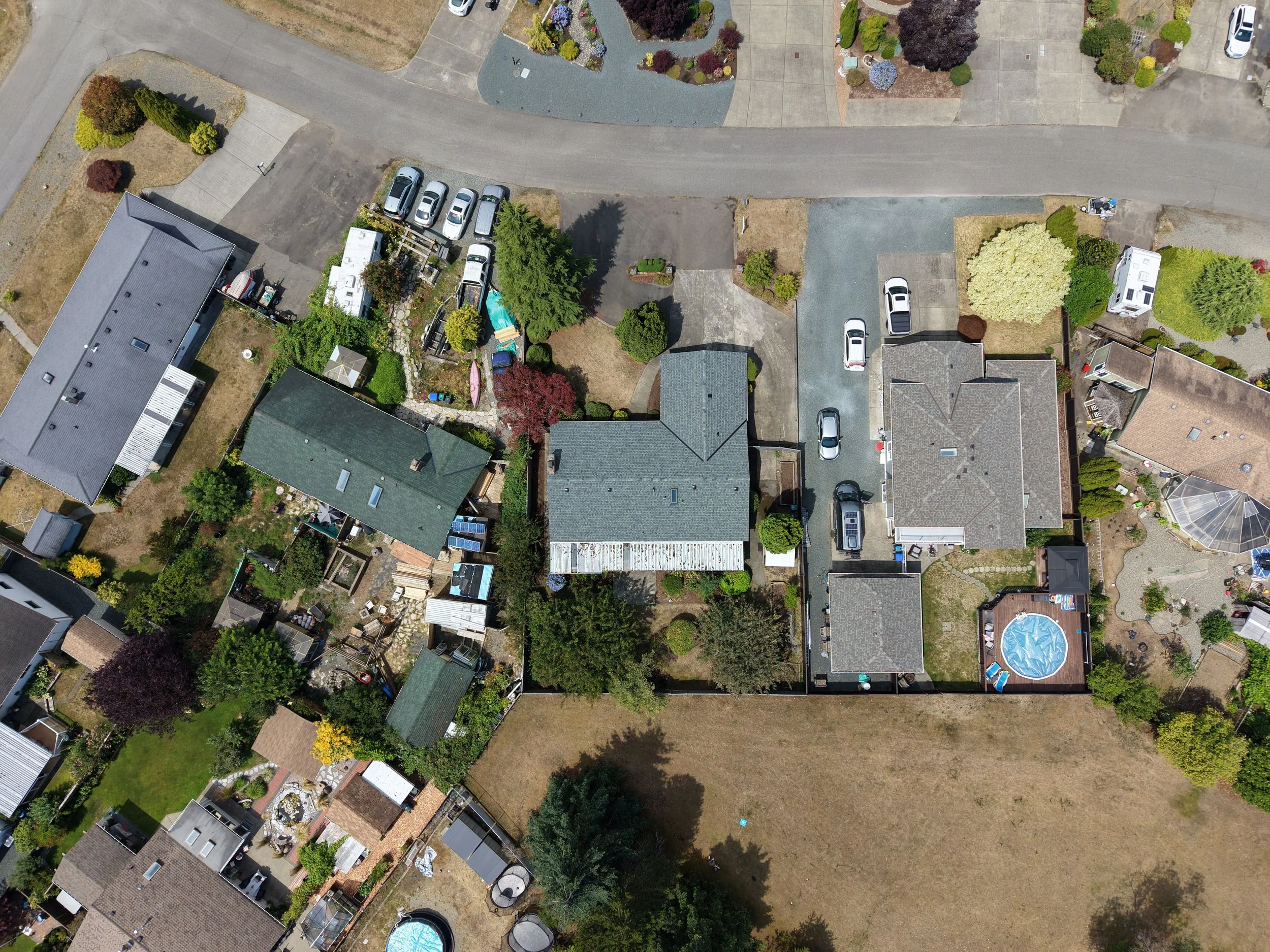 An aerial view of a residential neighborhood with houses, lush green trees, driveways, parked cars, gardens, and a backyard with a swimming pool.