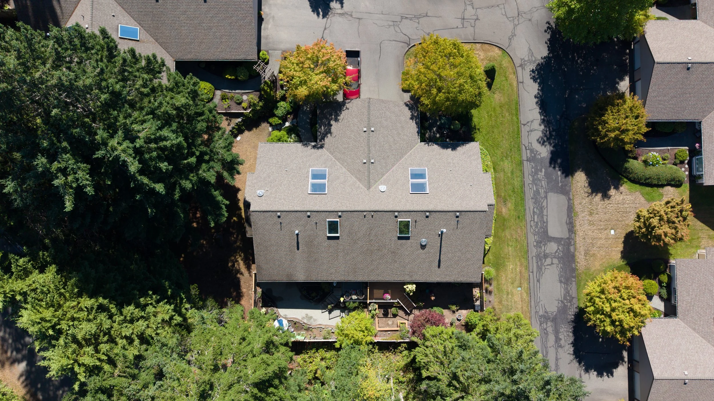 An aerial view of a residential neighborhood showing houses with grey roofs, surrounding trees, and a driveway with red and black vehicles parked.