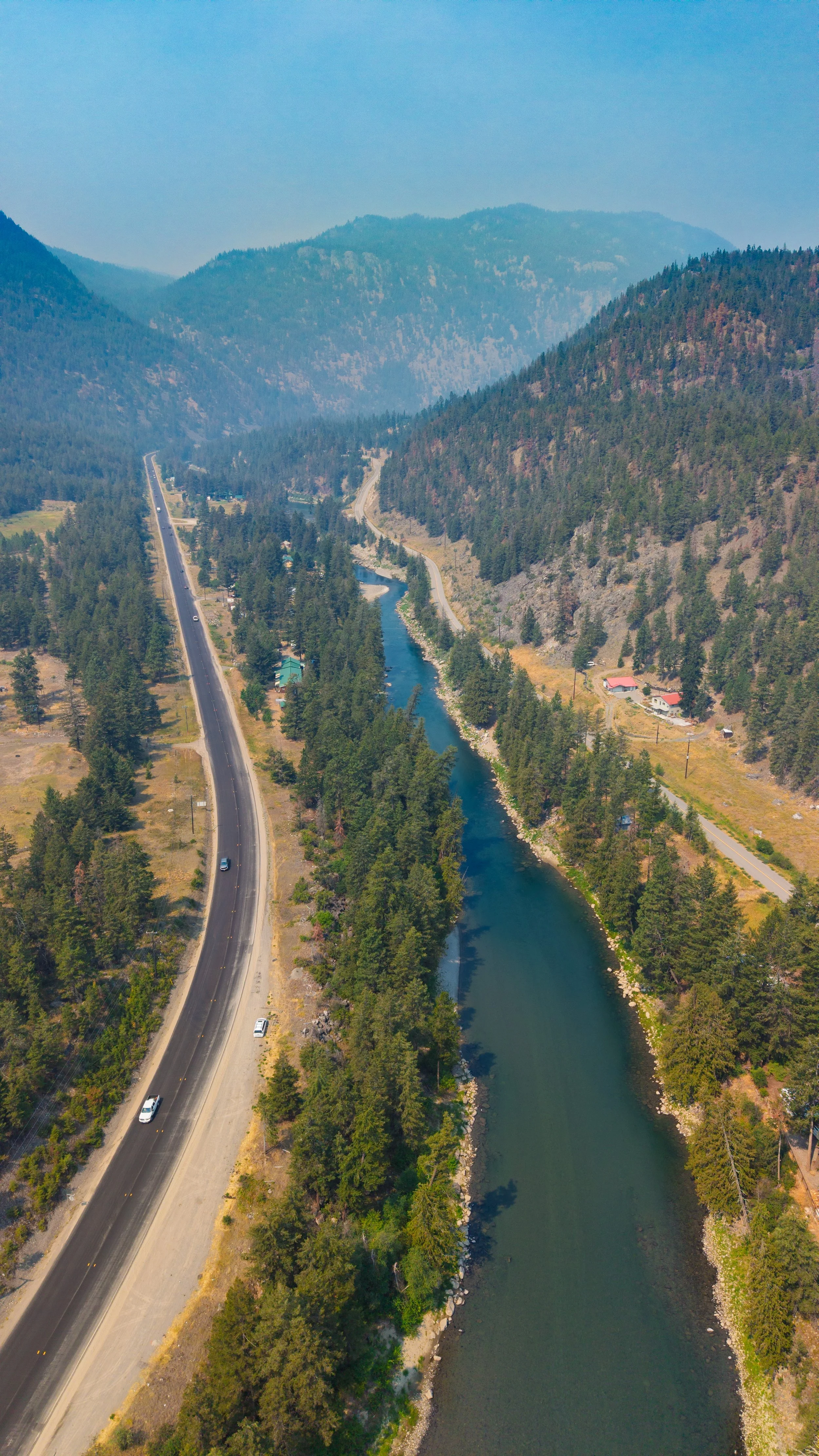 Aerial view of a winding river flowing through a forested valley with mountains in the background and a road running parallel to the river on the left side.