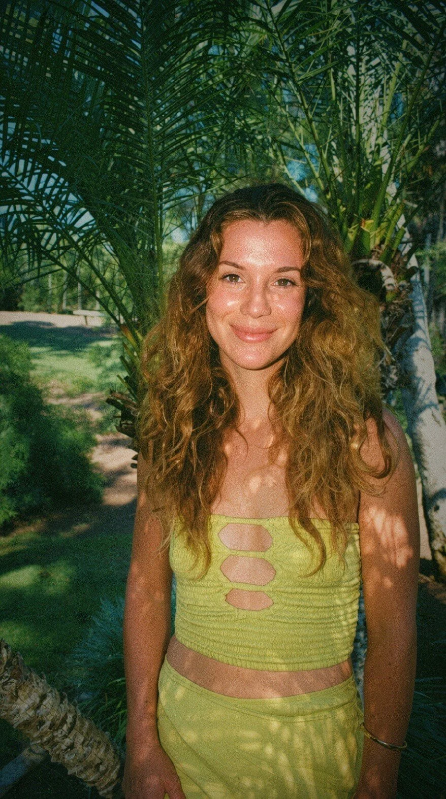 A young woman with long curly hair standing outdoors in front of green tropical plants, smiling at the camera, wearing a yellow cut-out top and matching skirt.