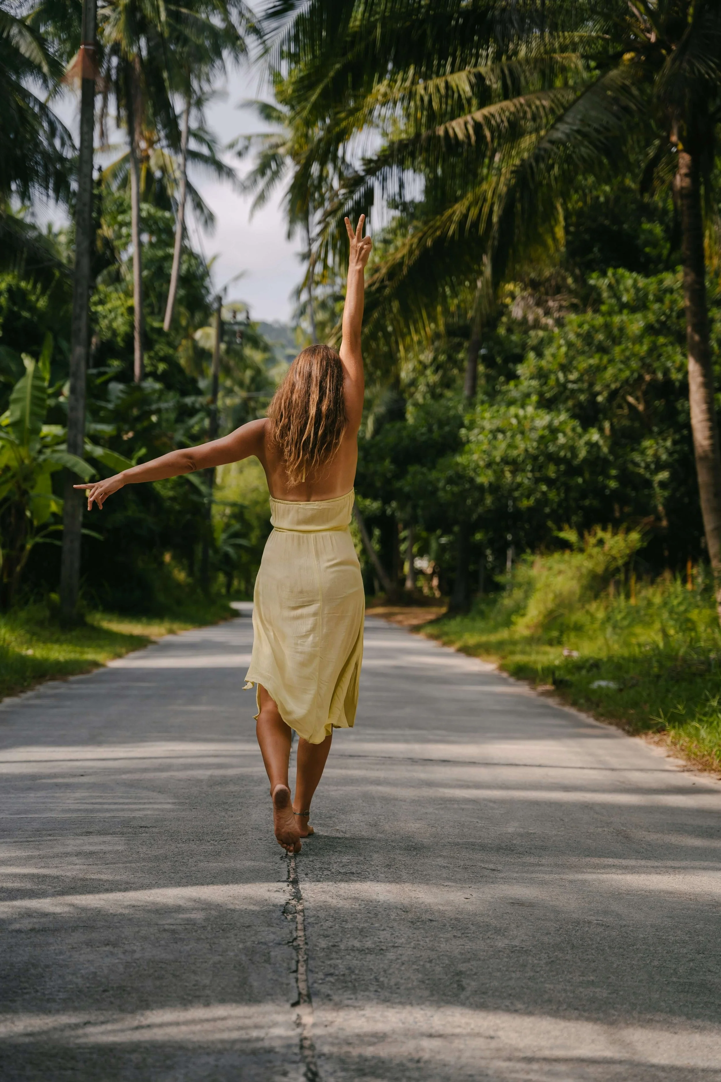 A woman walking barefoot on a rural road with dense trees and palm trees around, raising her right arm with two fingers in a peace sign and wearing a strapless yellow dress.