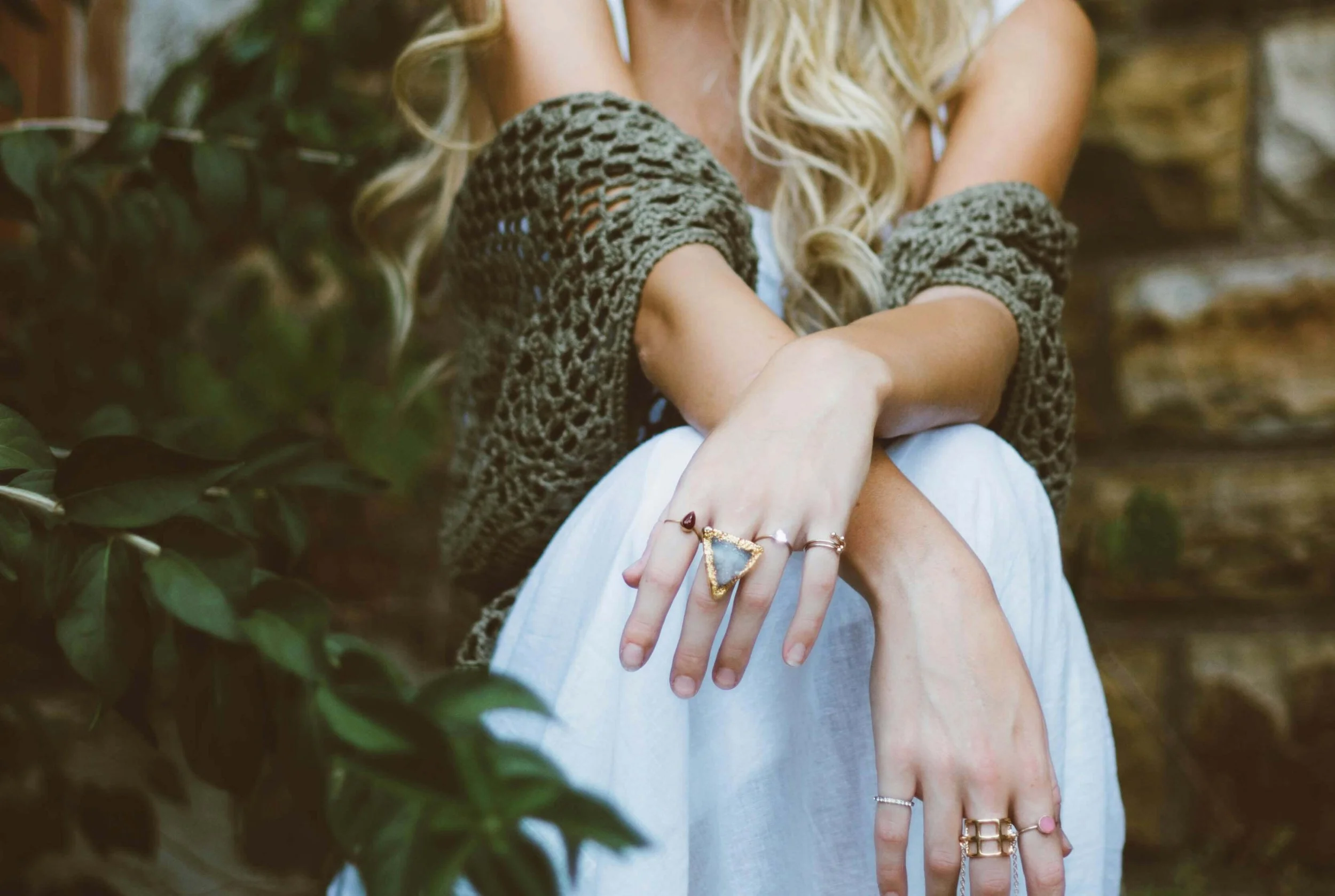 Person with long blonde hair wearing rings on fingers, a gray knitted cardigan, and a white dress, standing outdoors near green foliage.