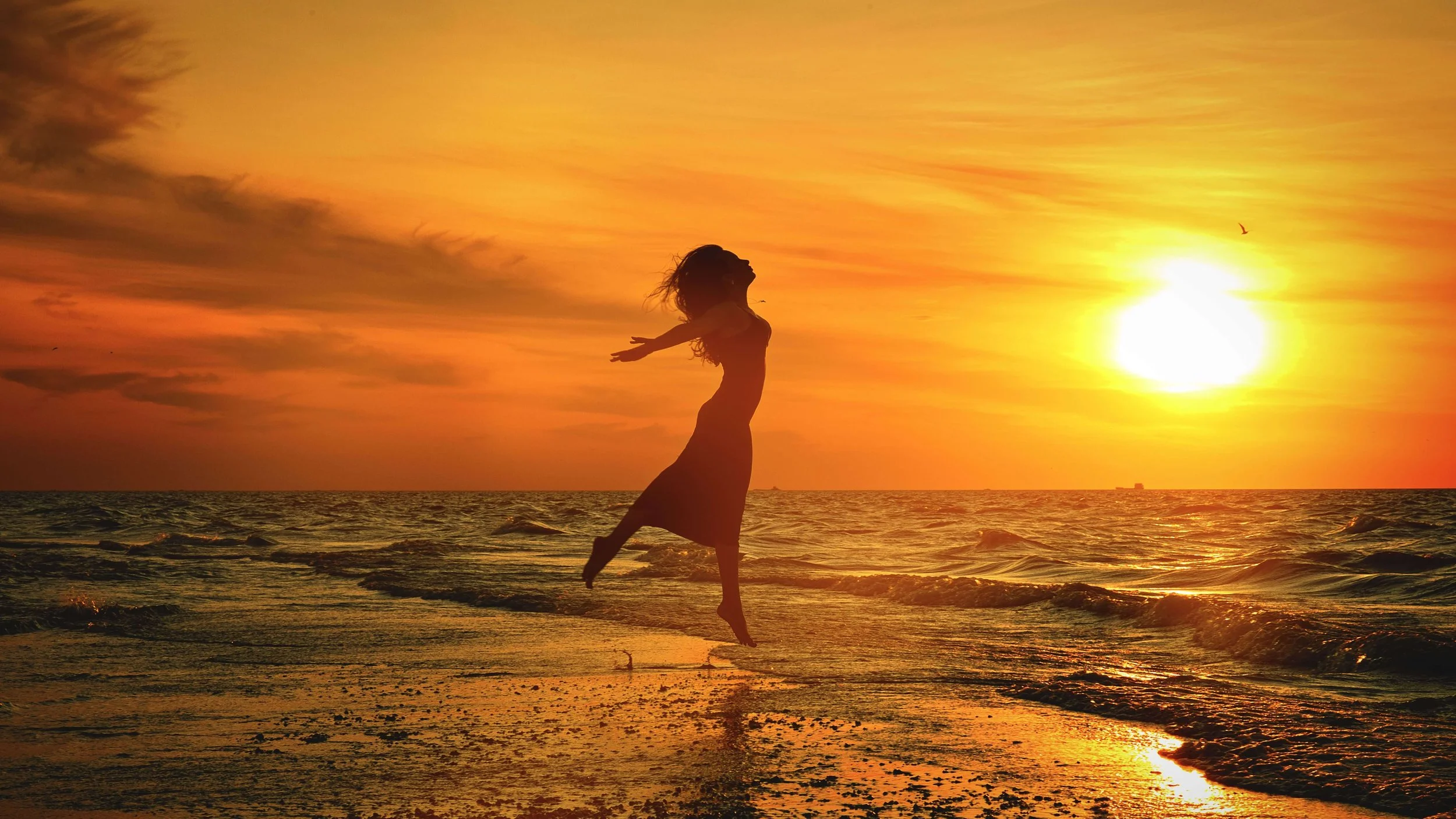 A silhouette of a woman jumping on the beach at sunset with the ocean and sky illuminated in orange and yellow hues.