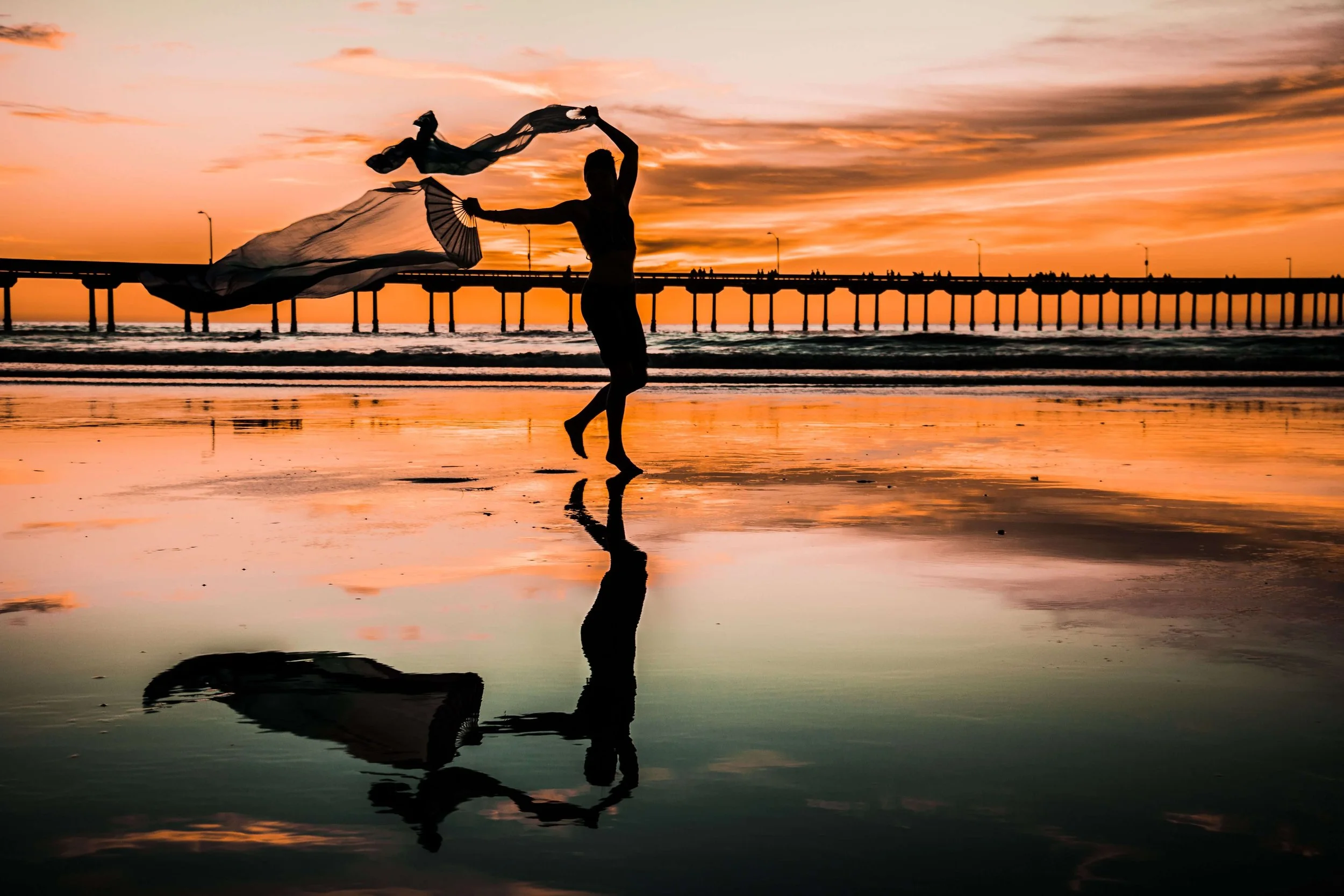 Person dancing on the beach during sunset, holding two fans and flowing fabric, with pier in the background and reflections on wet sand.