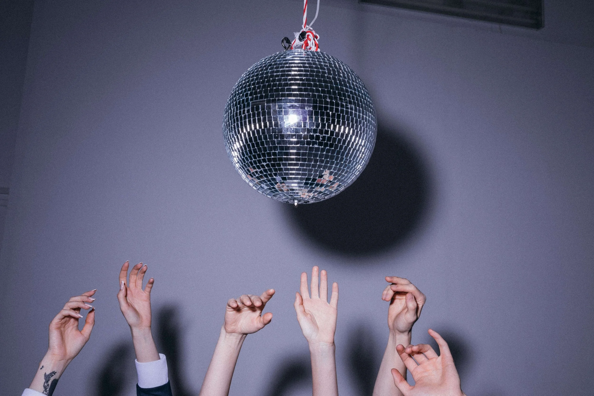 Multiple hands reaching towards a disco ball hanging from above against a gray wall.
