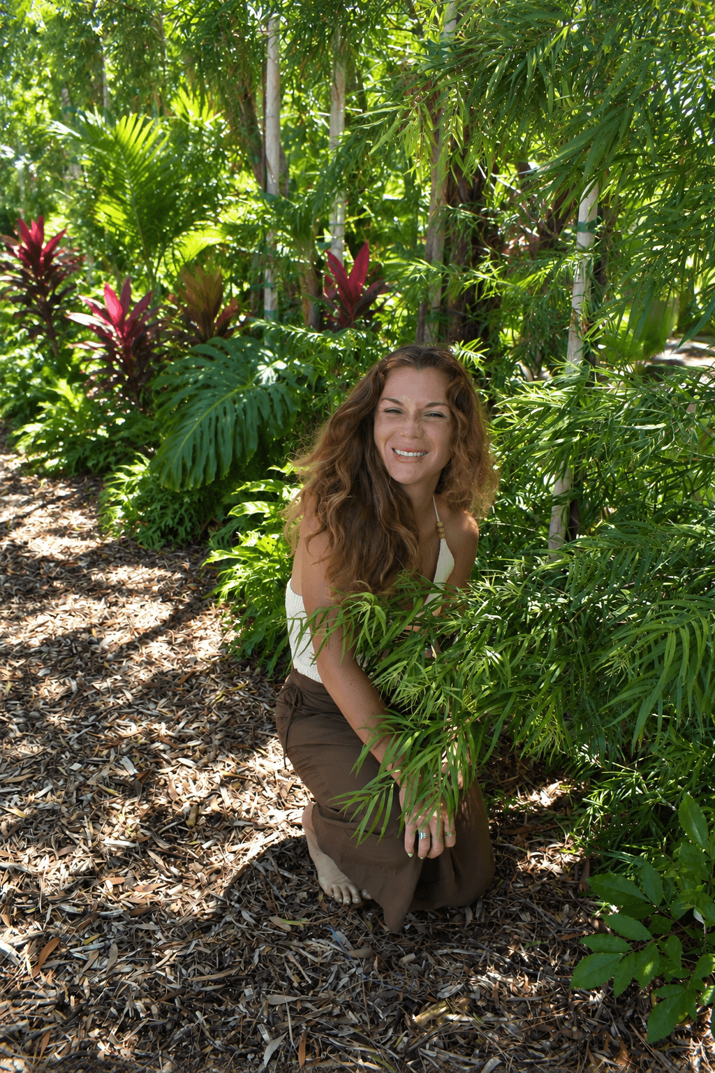 Woman with curly hair smiling and crouching among lush green tropical plants.