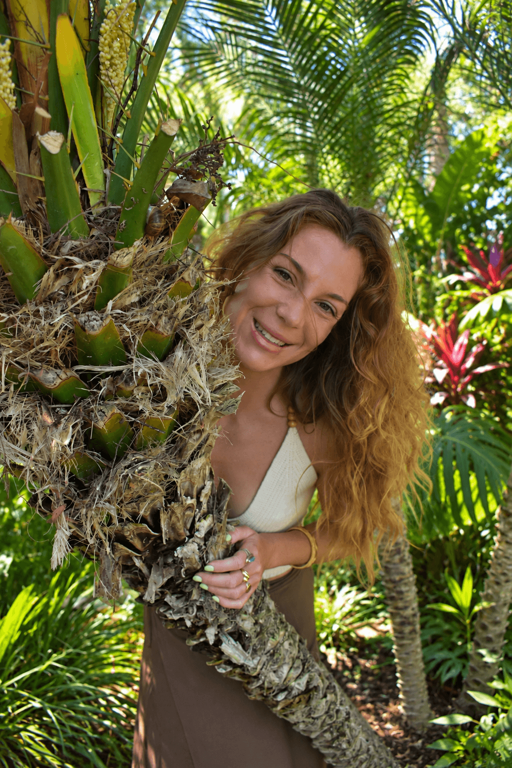 A woman with long curly hair leaning on a palm tree in a lush tropical garden.