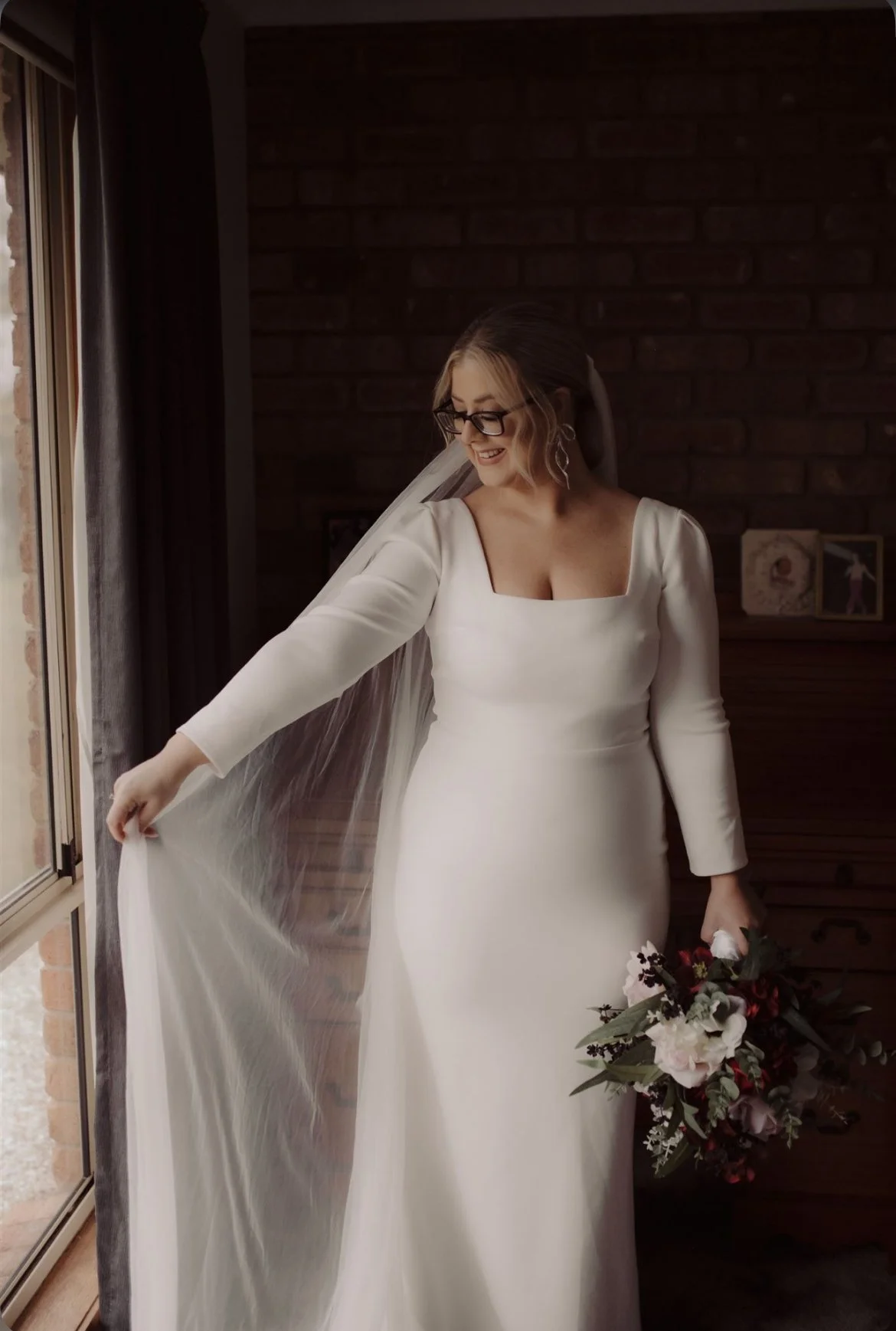 Bride in a white wedding dress with long sleeves, holding a bouquet of flowers, standing indoors near a window with dark brick wall background.