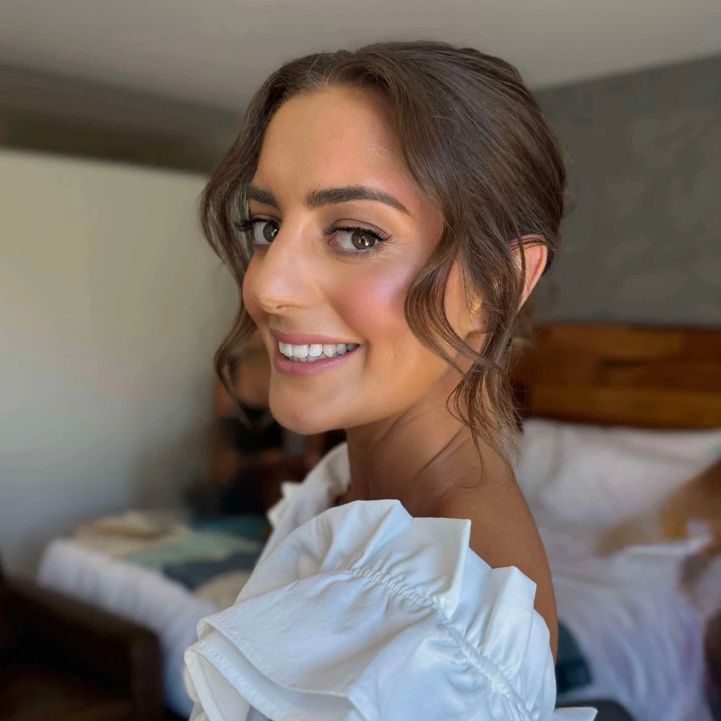 A smiling woman with short wavy brown hair and light makeup, wearing a white top with ruffled sleeves, in a bedroom with a bed and a woman in the background.