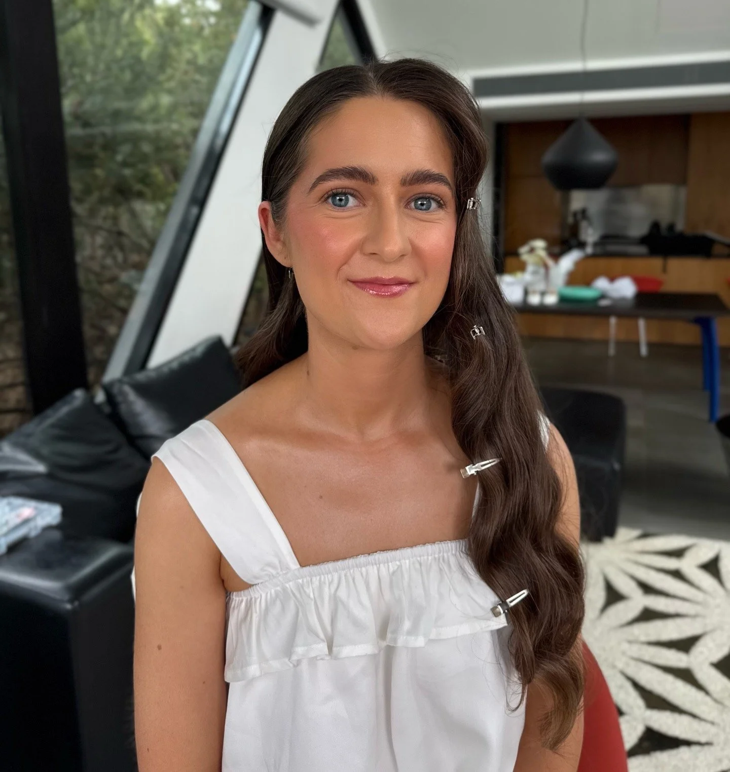A woman with long brown hair styled in waves and blue eyes, smiling, wearing a white sleeveless top with ruffle details, sitting in a modern living space with large windows, black leather furniture, and a patterned rug.