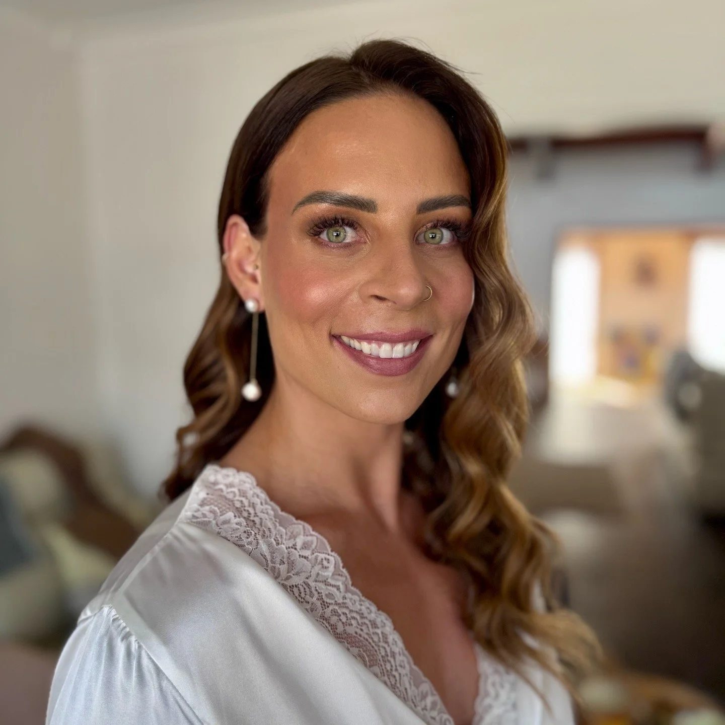 Close-up of a smiling woman with light skin, green eyes, wavy brown hair, wearing pearl earrings and a white satin robe with lace trim in a softly lit room.