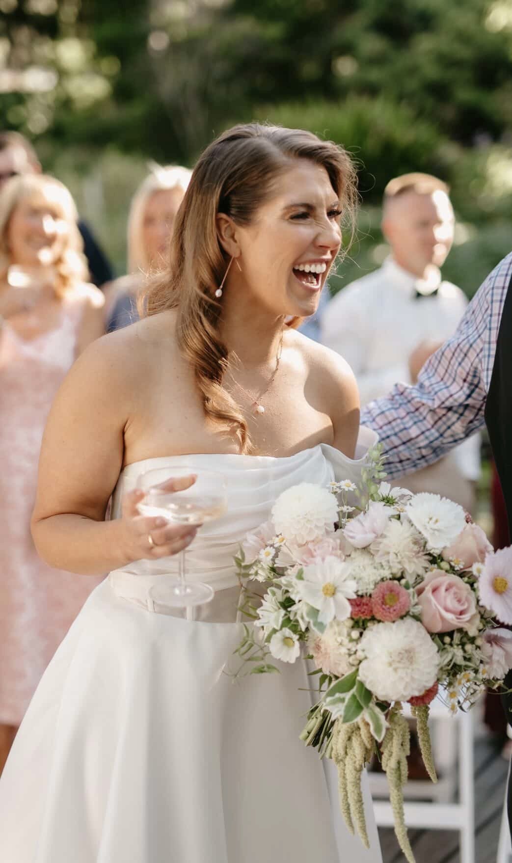 A bride with long brown hair in loose curls is holding a bouquet of white and pink flowers at an outdoor wedding. She is wearing a strapless white wedding dress and is laughing while holding a glass of wine, with guests smiling in the background.