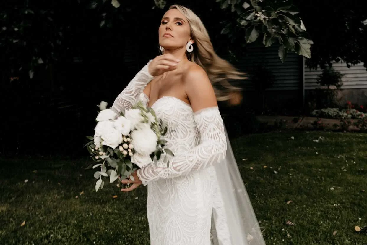 A woman wearing a white lace off-shoulder wedding dress, holding a bouquet of white flowers and greenery, standing outdoors with trees and grass in the background.