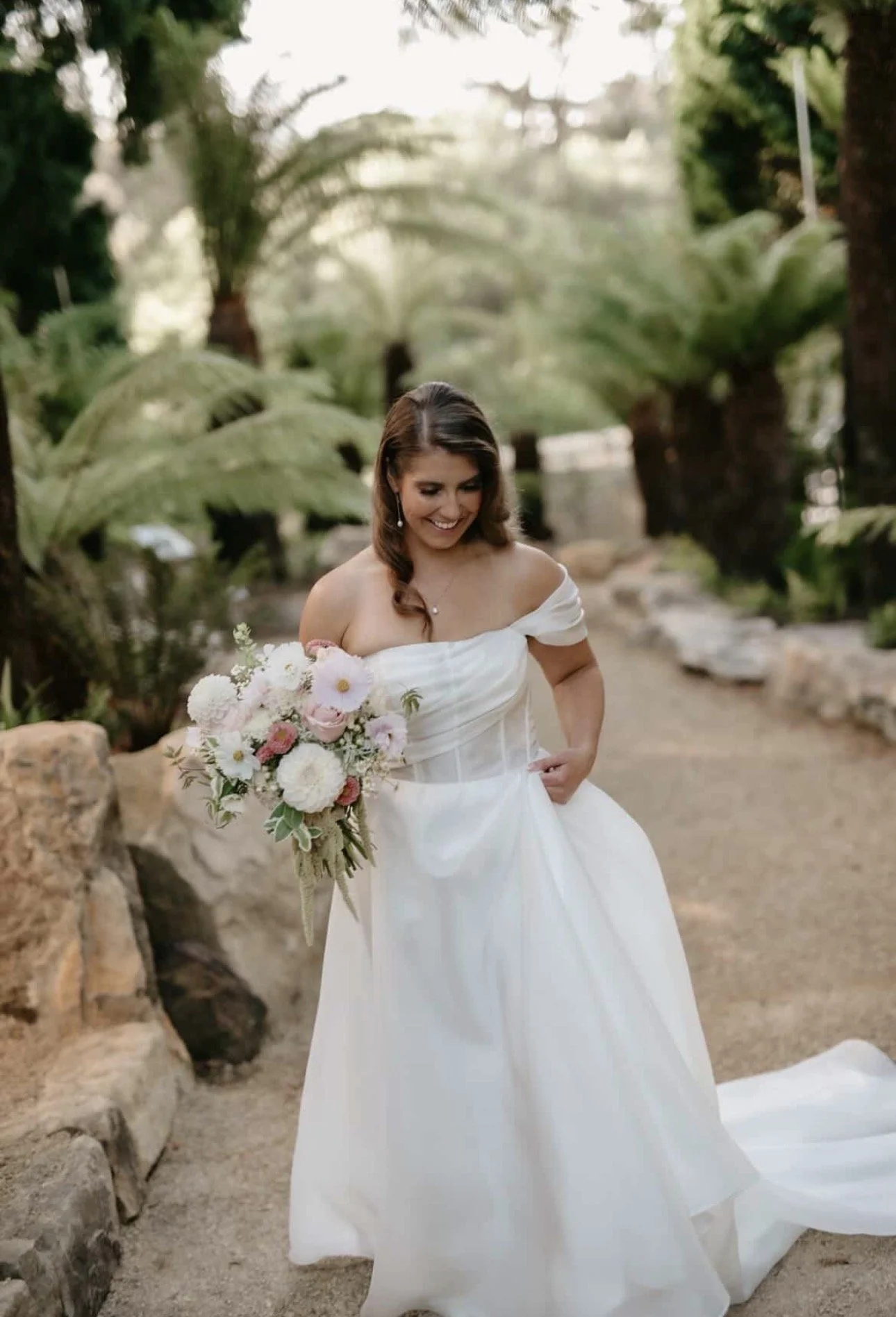 A woman in a white off-the-shoulder wedding gown holding a bouquet of white and pink flowers, smiling as she walks outdoors in a lush garden with trees and large green plants around her.