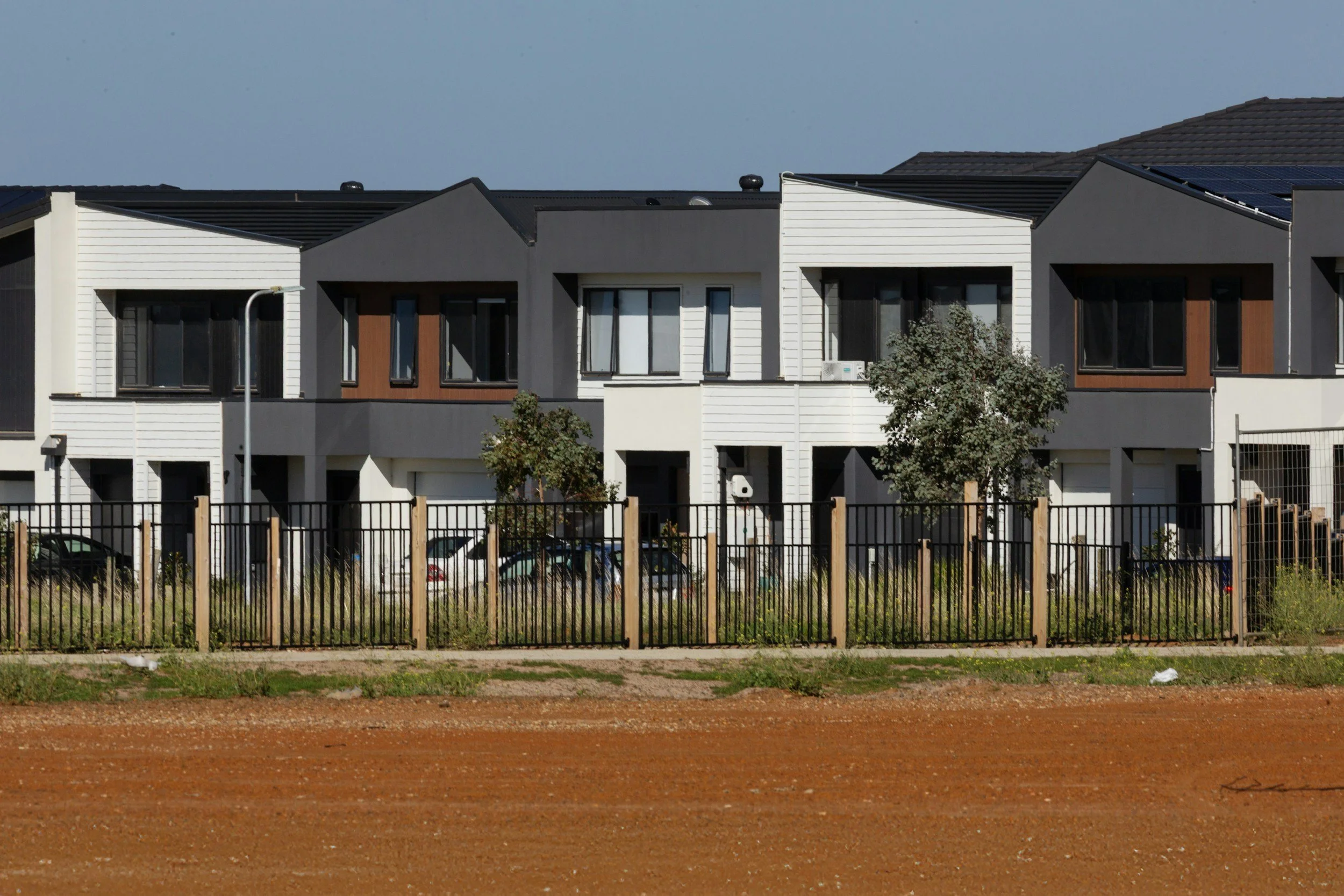 Modern multi-story residential building with various window styles, a white and gray exterior, and a wooden accent wall. There is a black metal fence in the foreground, some trees, parked cars, and a clear blue sky.