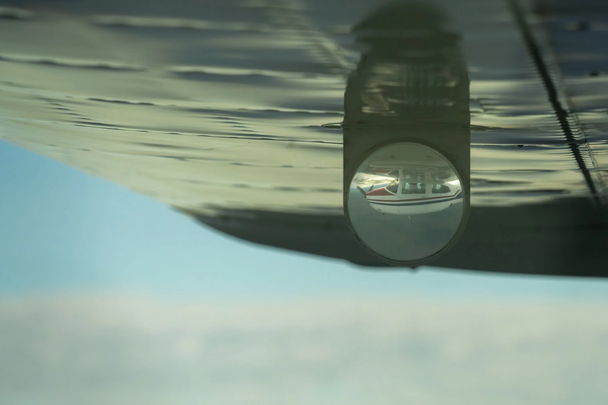 The image shows an aircraft flying upside down, with its underbelly visible from below. The aircraft has a streamlined body with a cockpit window and an attached antenna or wire visible on top. The sky in the background is clear and blue.