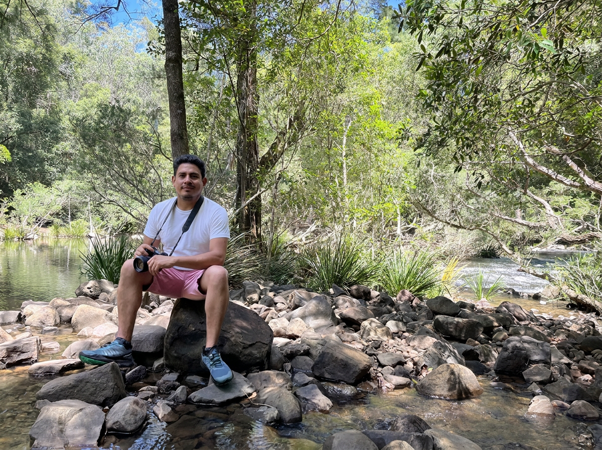 A man with a camera sits on a rock by a river surrounded by trees and foliage in a forest.