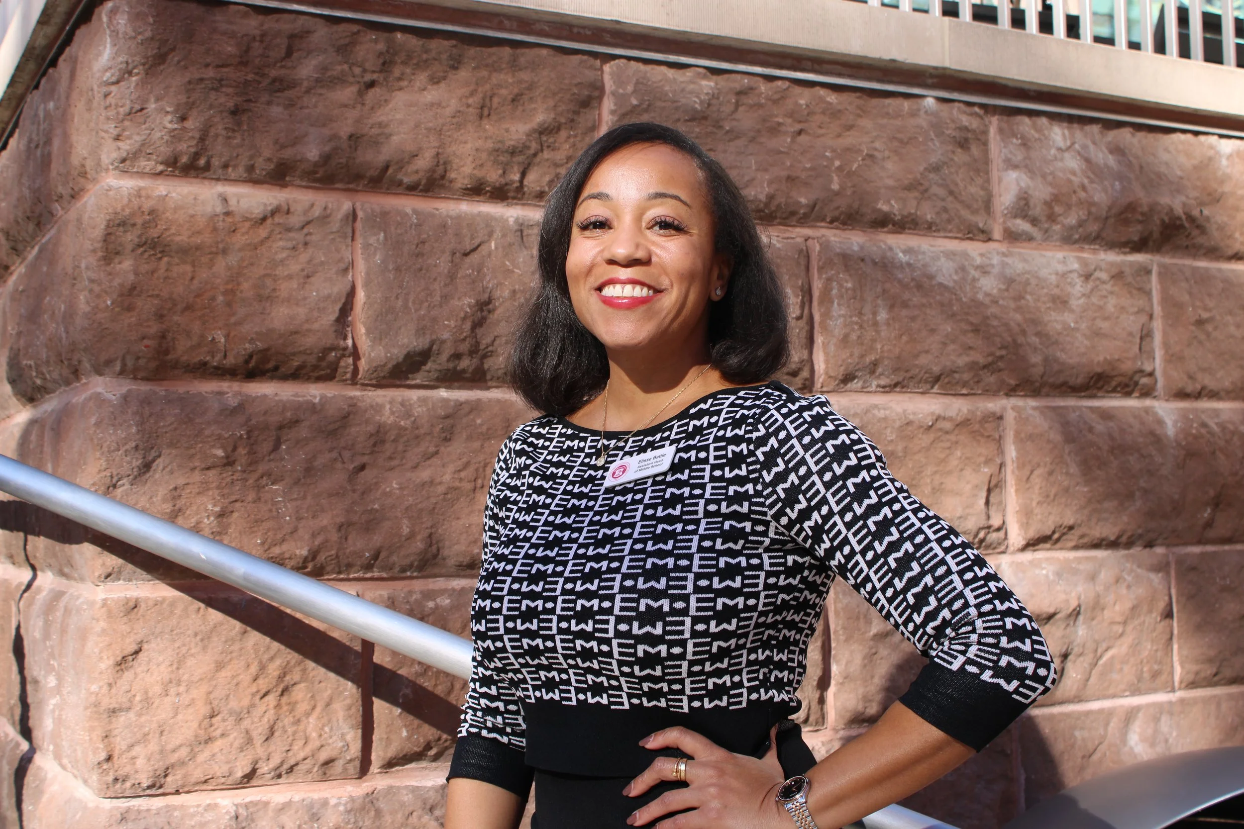 A woman with shoulder-length black hair smiling at the camera, wearing a black and white patterned top, standing outdoors in front of a stone building with a metal railing.