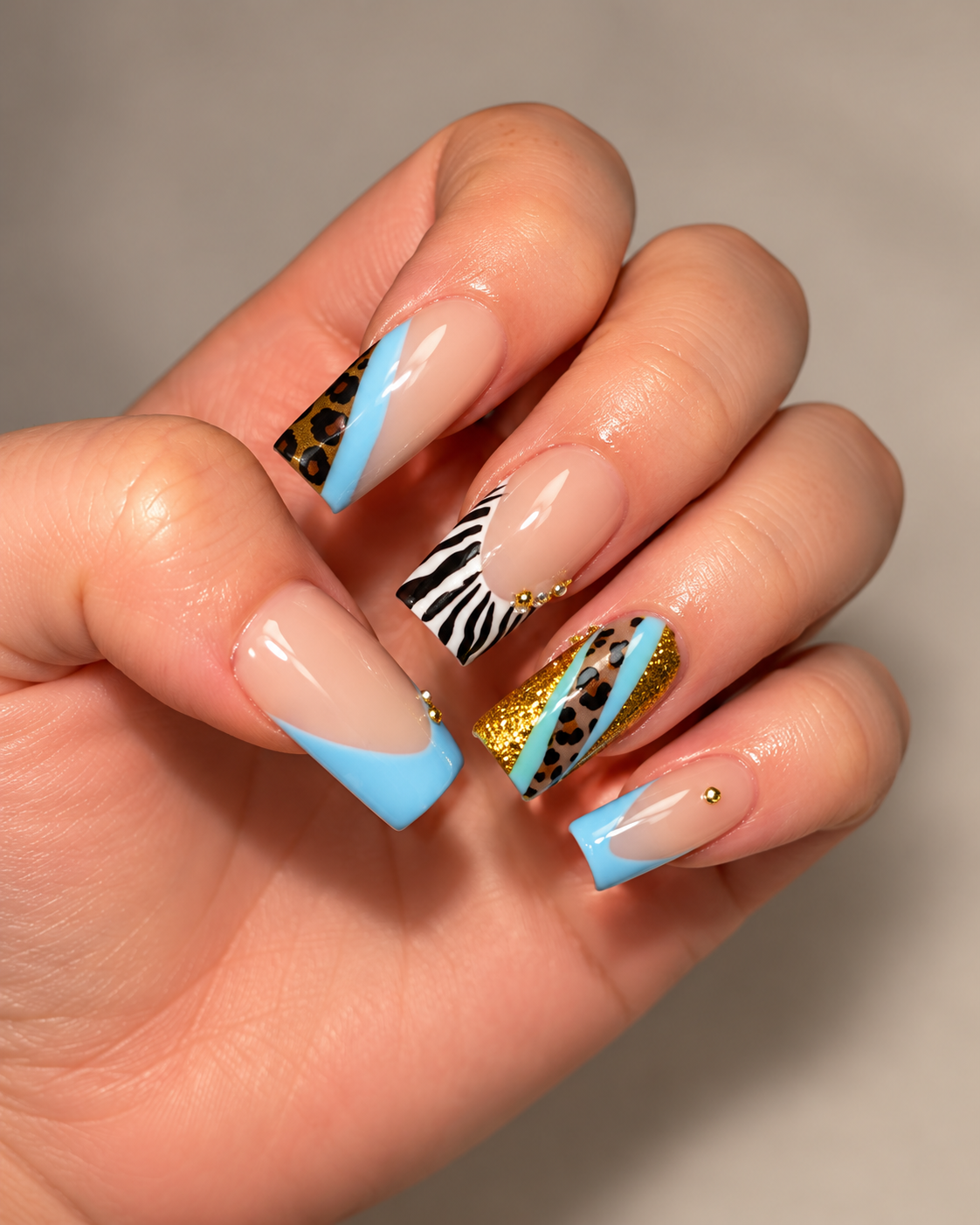 Close-up of a hand with decorated acrylic nails featuring animal print, glitter, and geometric patterns in blue, black and white, with small gold embellishments.