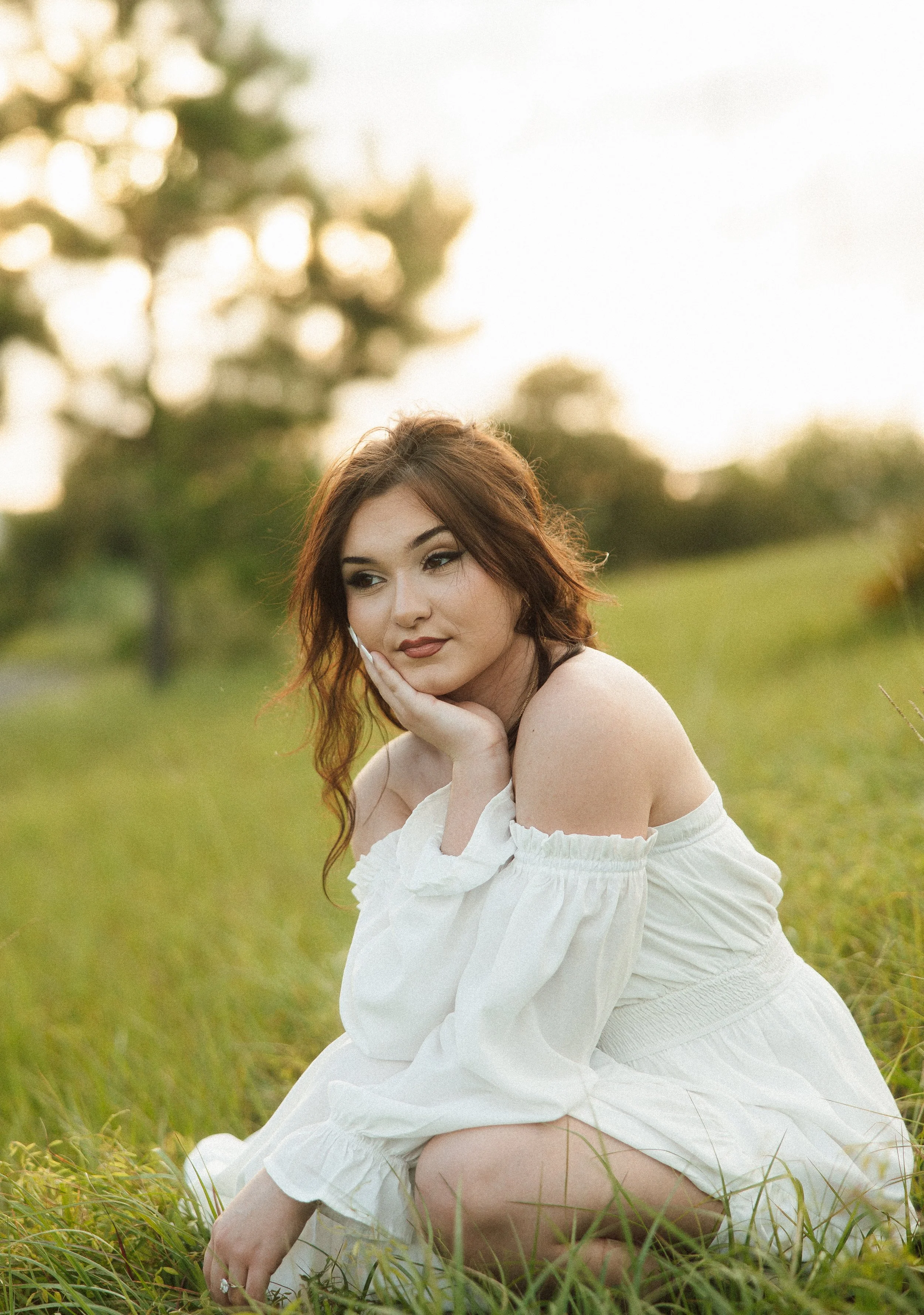 A young woman with auburn hair and makeup, wearing a white off-shoulder dress, sits on grass outdoors with trees in the background during golden hour.