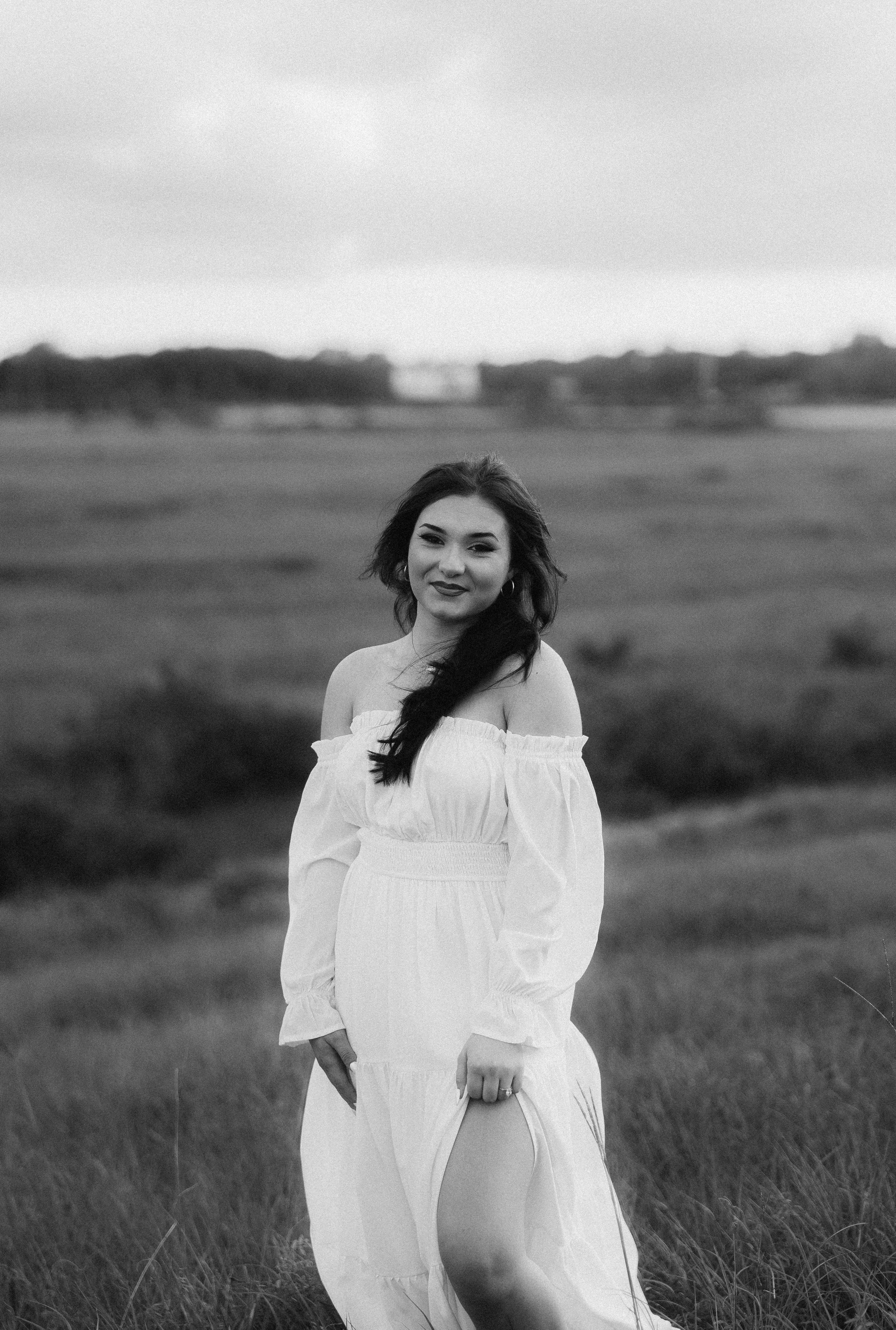 A black and white photo of a woman standing in a grassy field with trees in the background, wearing an off-the-shoulder white dress, with long dark hair, smiling, looking at the camera.