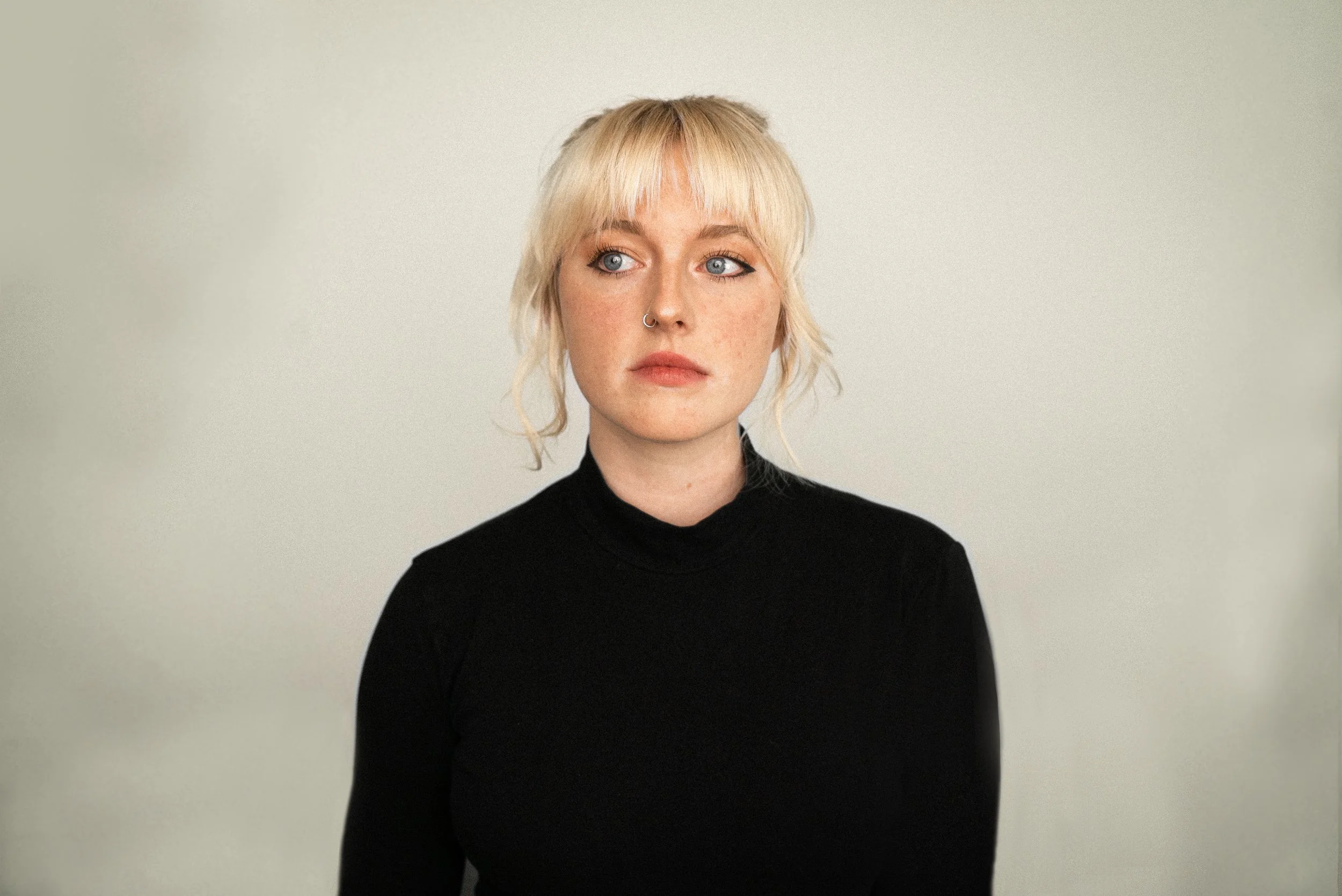 A woman with platinum blonde hair and blue eyes wearing a black turtleneck sweater, standing against a plain light-colored background.