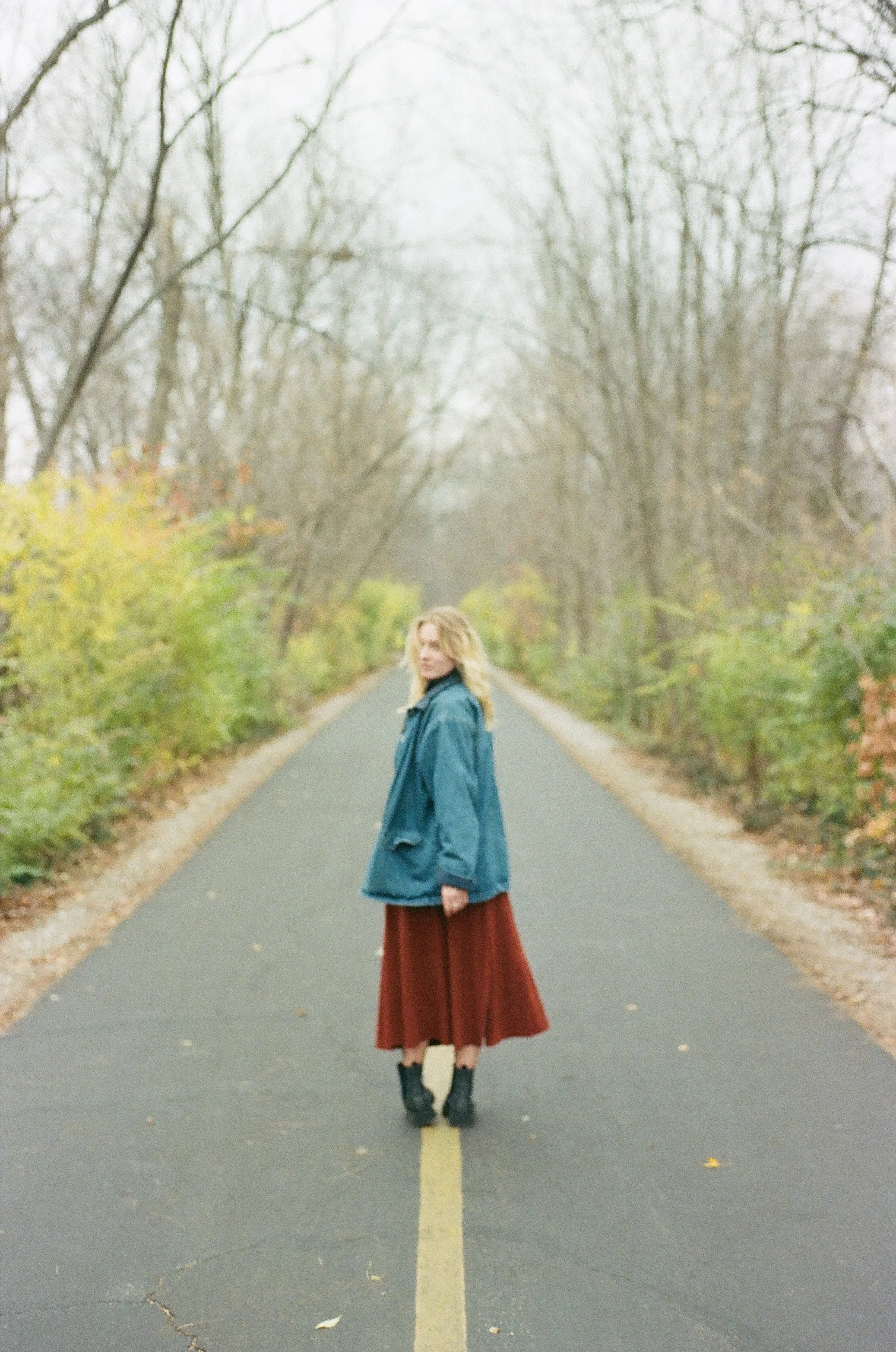 Woman with blonde hair standing in the middle of a paved road surrounded by trees with autumn foliage.