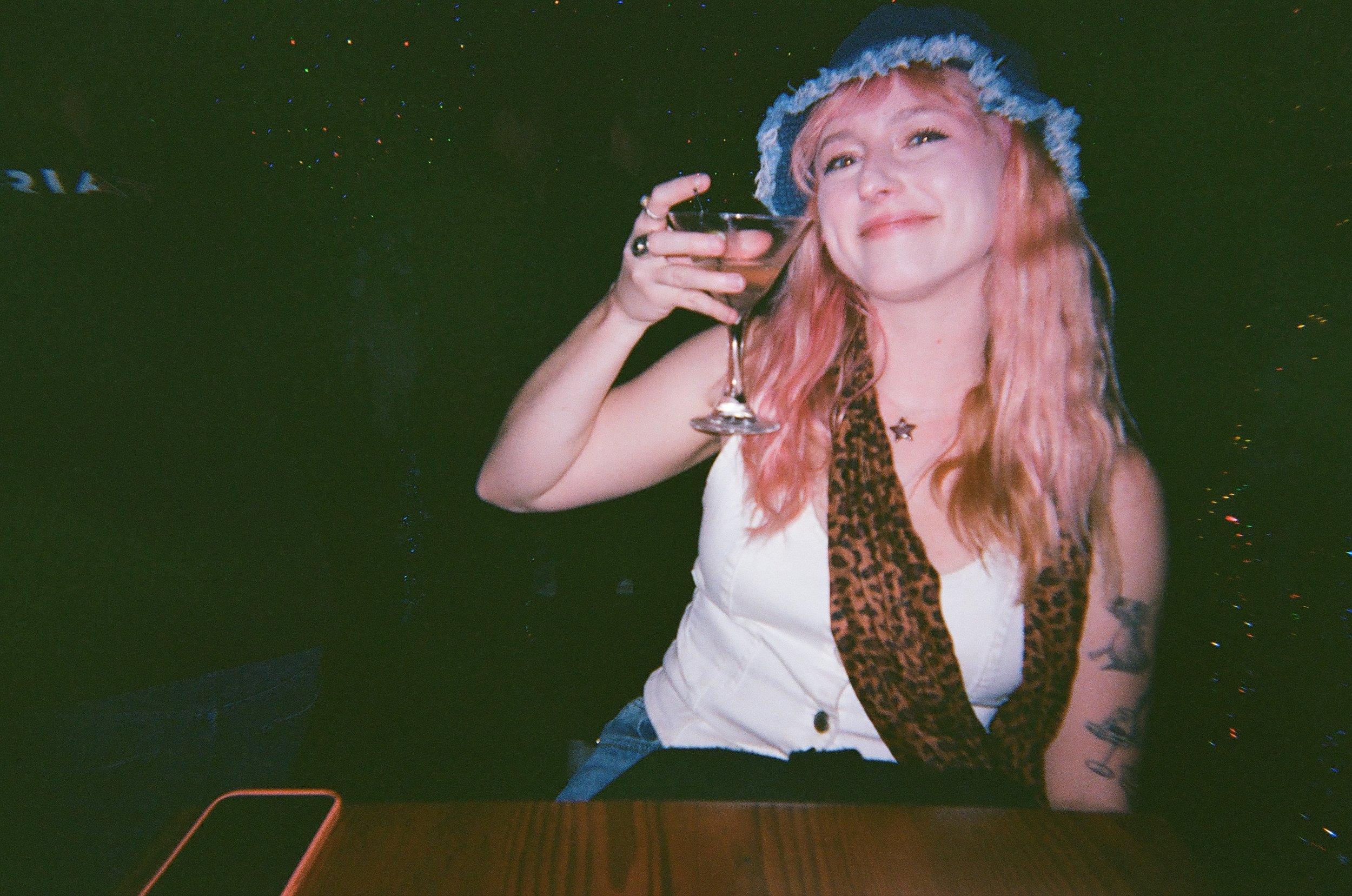 Woman with pink hair wearing a blue hat, holding a glass of drink, smiling, sitting at a table with a dark background with colorful lights.