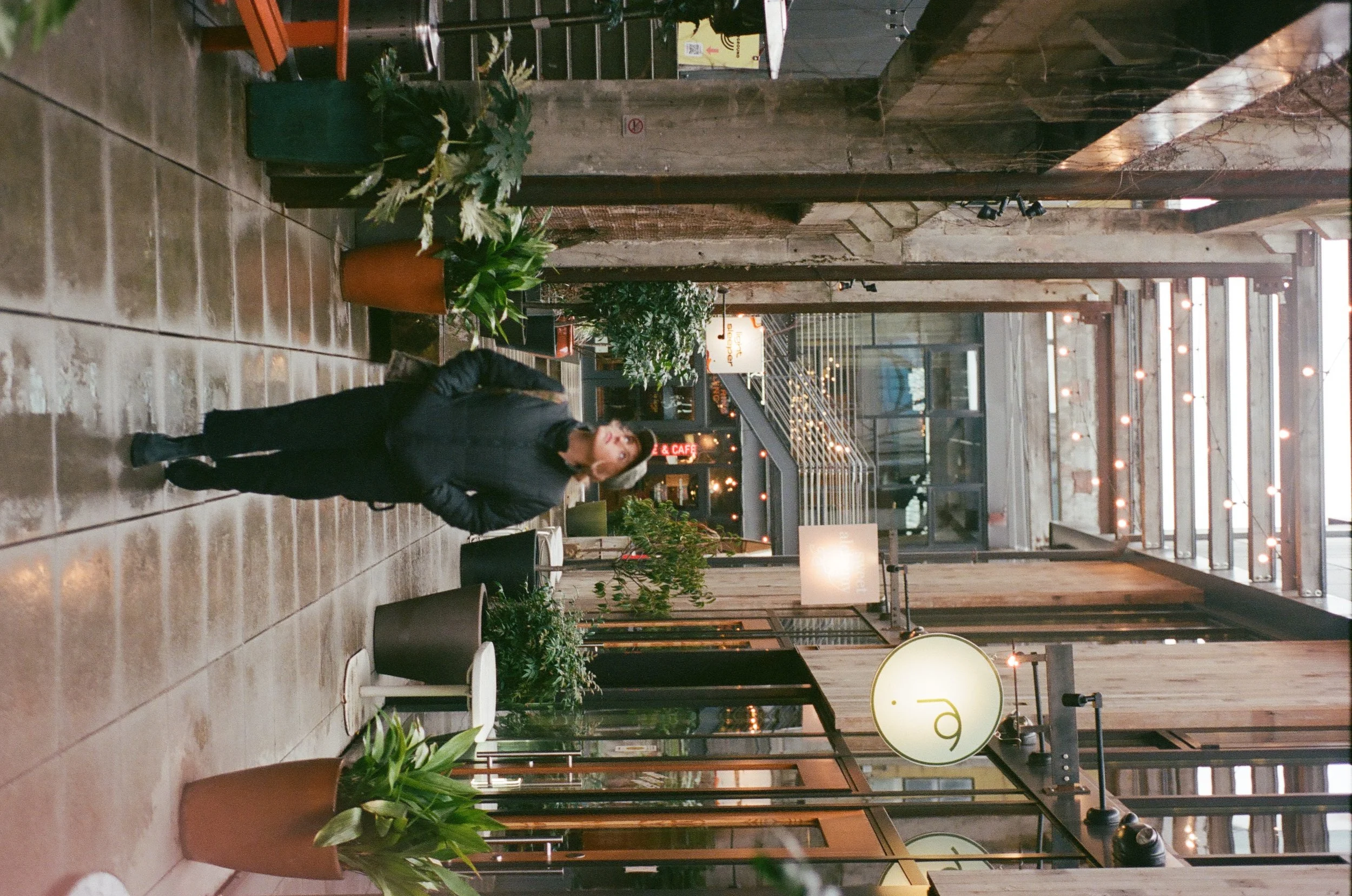 A woman walking through an indoor space with potted plants, wooden panels, and warm lighting, possibly a cafe or restaurant entrance.