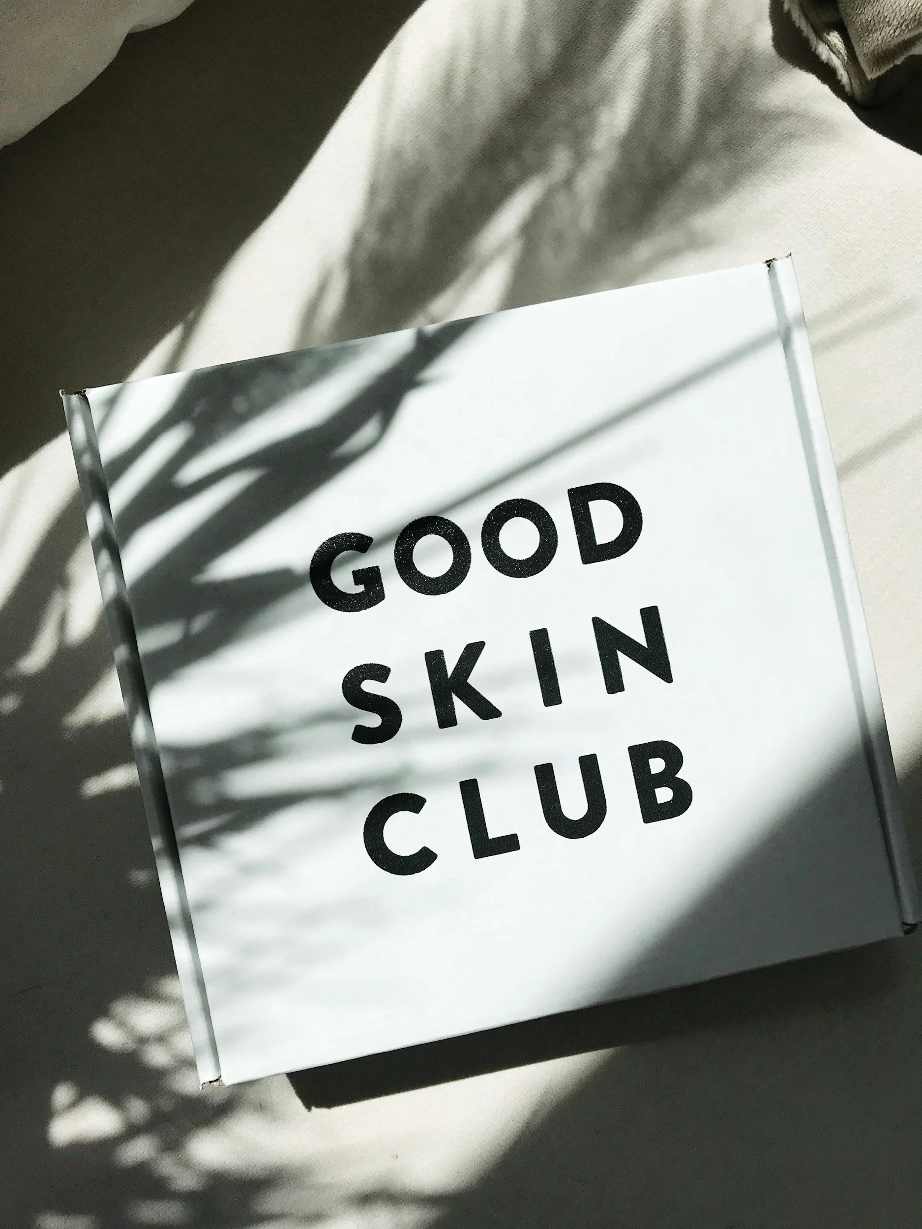 A white sign with black text that reads 'Good Skin Club' sits on a dark surface, partially illuminated by sunlight casting shadows.