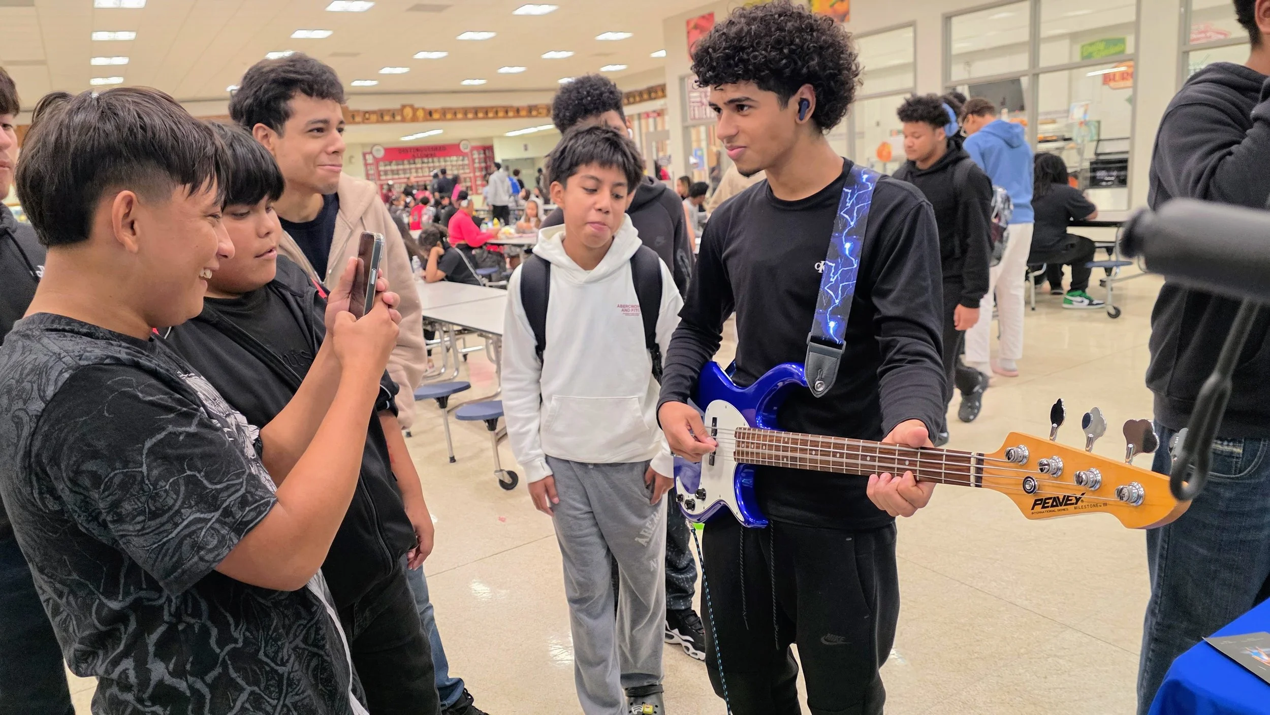 A group of boys in a school cafeteria, with one boy playing an electric guitar and others watching him and recording with a phone.