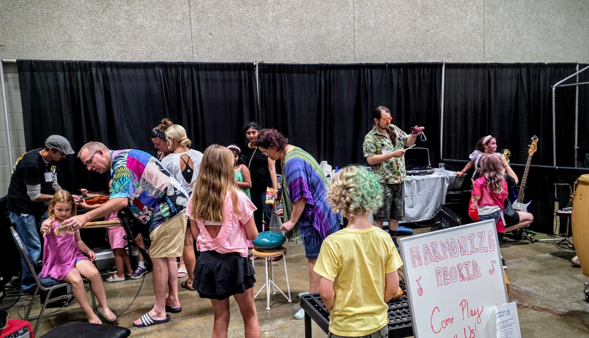 A diverse group of children and adults gathered at an indoor event with a black curtain backdrop. Some people are playing instruments and others are observing the activities. There is a sign that reads 'HARMONIZE' and another partially visible sign with the words 'PEOPLE' and 'Come Play with Us!'.