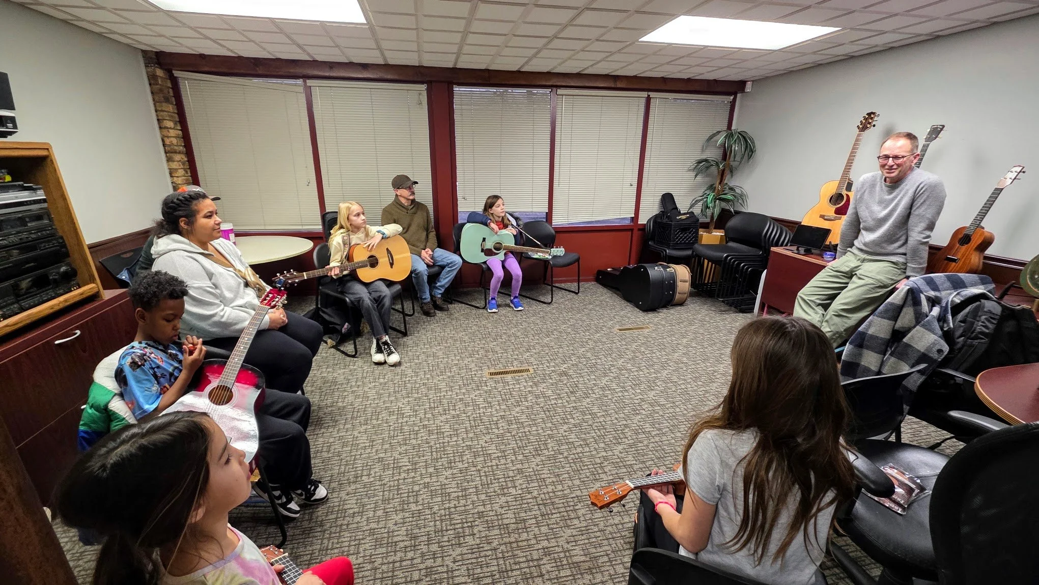 Children and adults in a music room, sitting with guitars, listening to a man at the front who appears to be instructing or speaking. The room has large windows with blinds, a potted plant, and guitar cases.