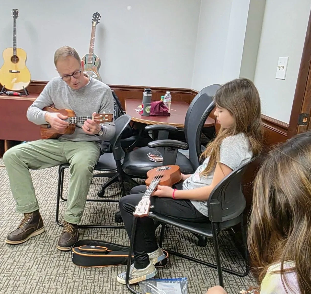 A man and a young girl are sitting in chairs in a room playing guitars. The man is playing an acoustic guitar, and the girl is holding a smaller guitar on her lap. There are additional guitars hanging on the wall behind them. The room has a carpeted floor, a table with water bottles, a backpack, and other items on it.