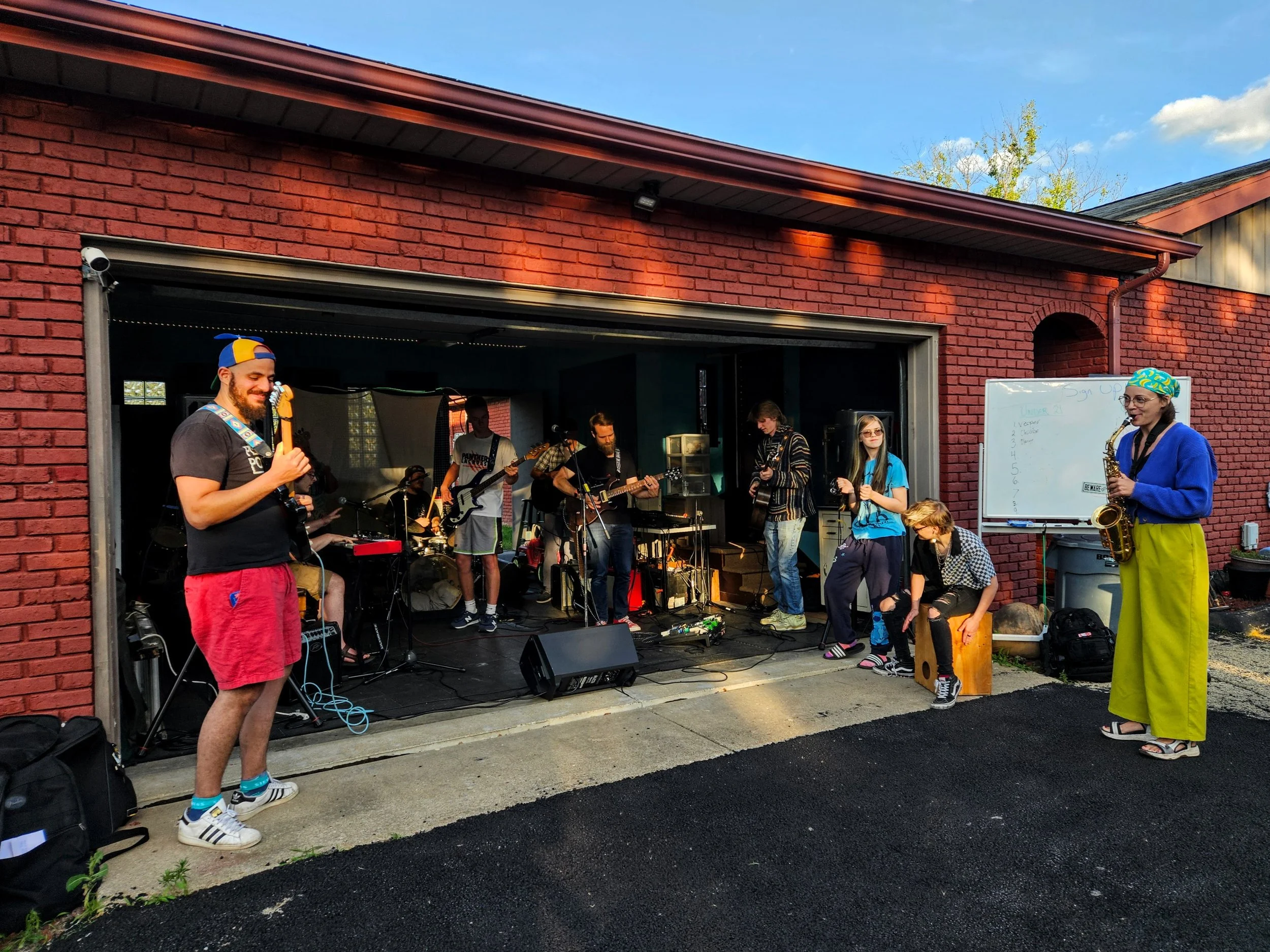 A group of people playing music on a stage set up in a garage with a brick wall behind them. Some are playing instruments, while others are standing or sitting nearby, enjoying the outdoor performance.