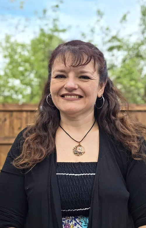 A woman with curly brown hair smiling outdoors, wearing hoop earrings, a black cardigan, and a necklace with a decorative pendant, with a wooden fence and green trees in the background.