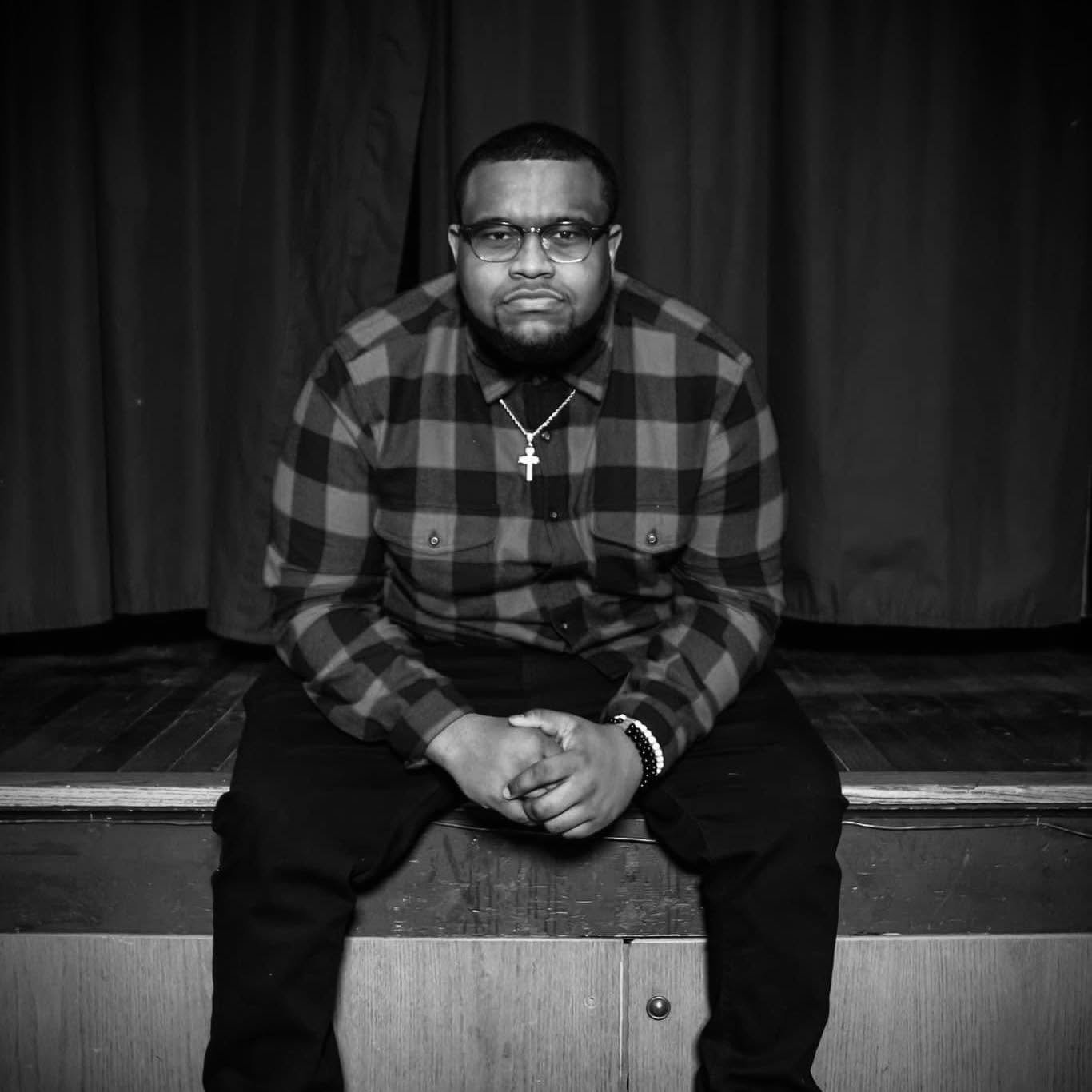 A man sitting on a stage, wearing glasses, a plaid shirt, and a cross necklace, with dark curtains in the background.