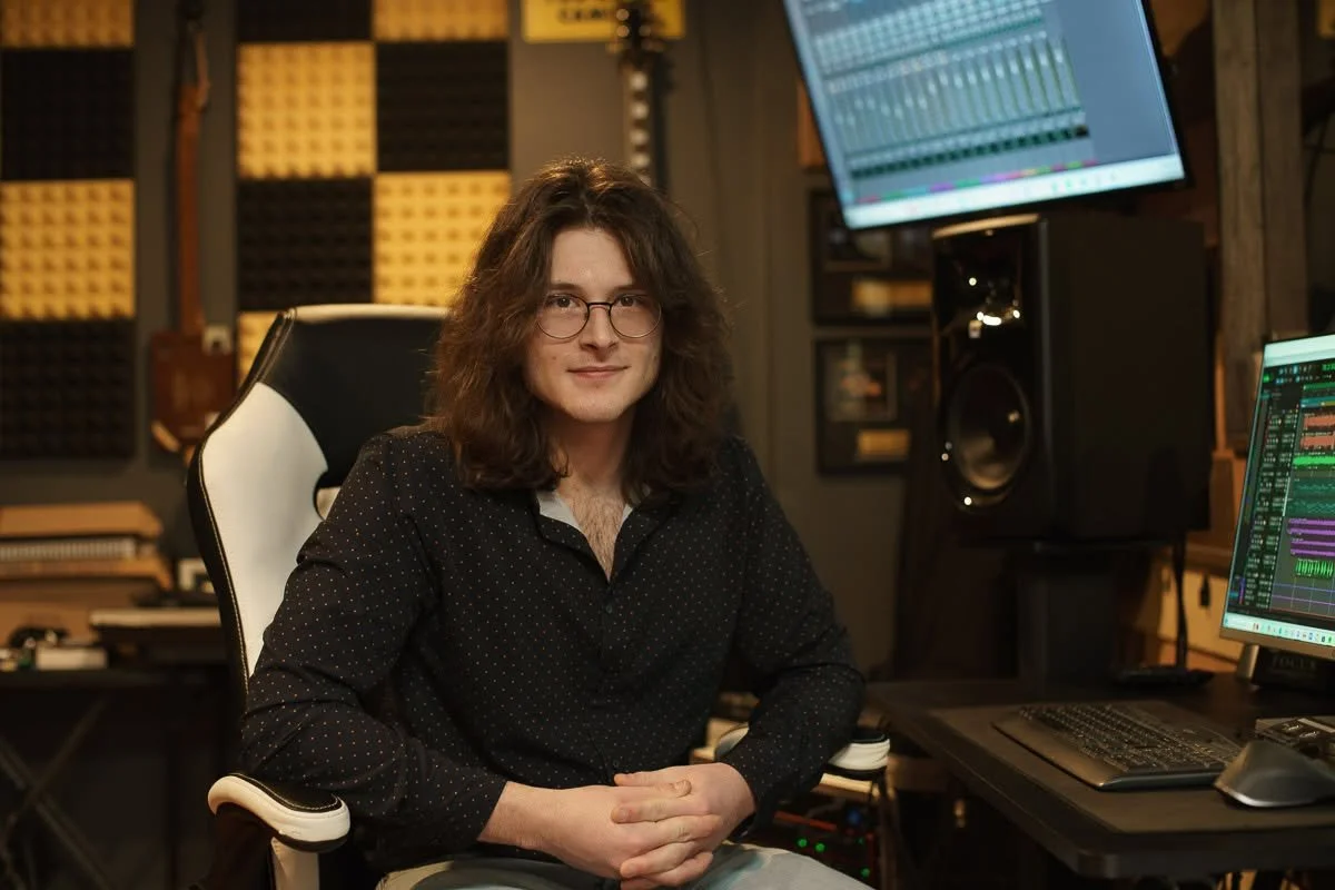 A person with long, wavy brown hair and glasses sitting in a recording or music production studio with computer screens displaying audio editing software.