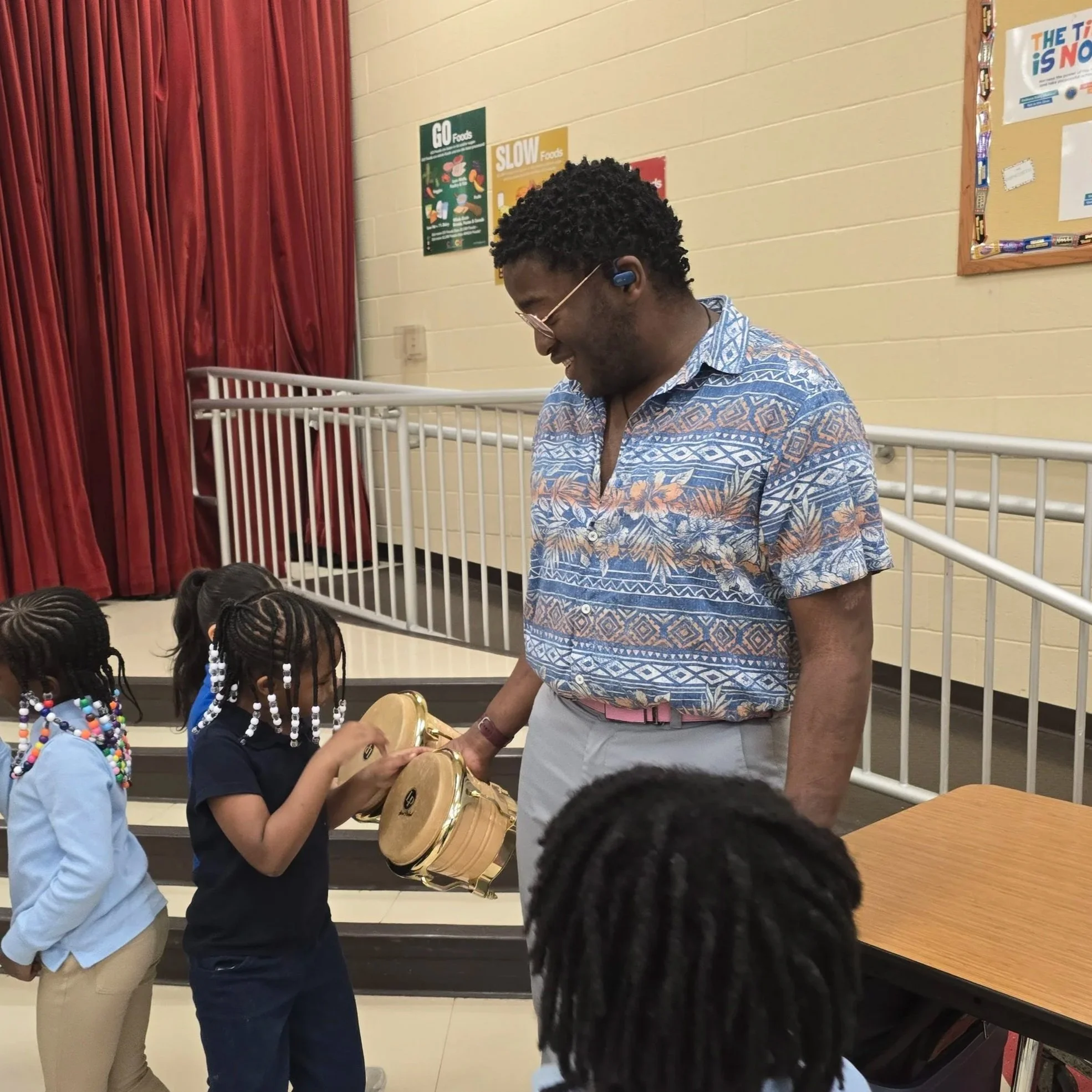 A man with curly hair, glasses, and a blue patterned shirt smiling while interacting with children. The children at the front have braided hair with colorful beads, and one child is holding a tambourine. The background includes a red curtain, beige wall, and educational posters.