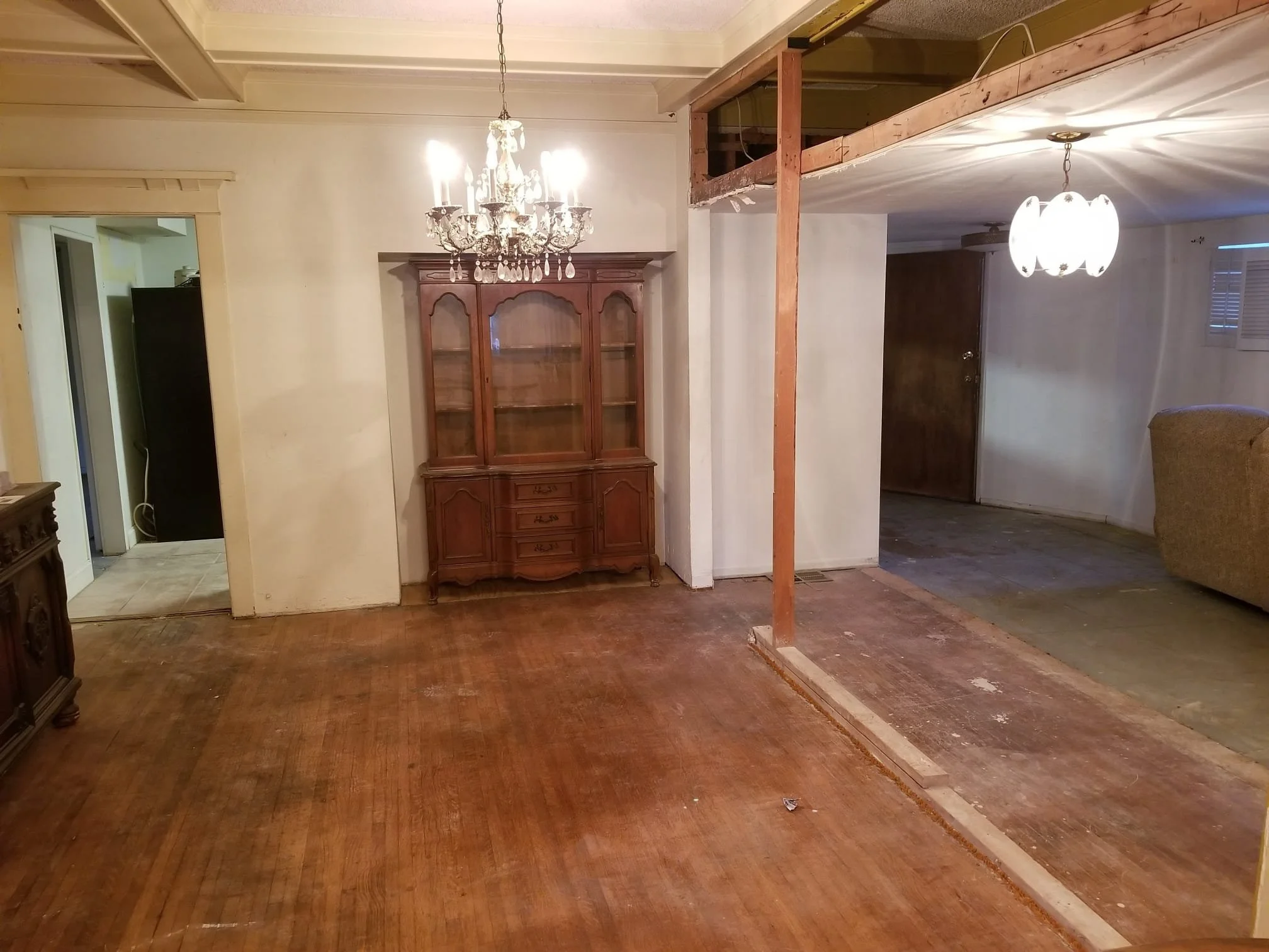 Living room undergoing renovation with exposed wall studs, chandelier, cabinet, and hardwood flooring.