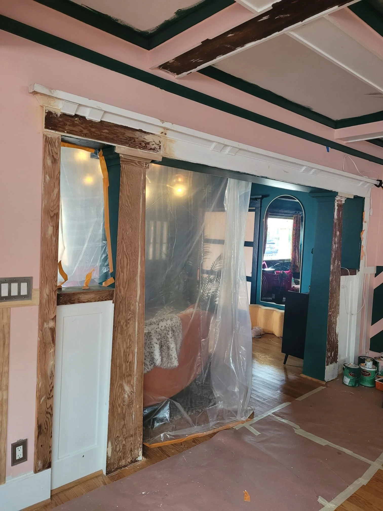 Interior of a room under renovation with exposed wooden framing around a doorway, plastic sheets blocking the doorway, and paint cans on the floor. The ceiling has a built-in design with pink and dark green accents, and the room includes a window wit